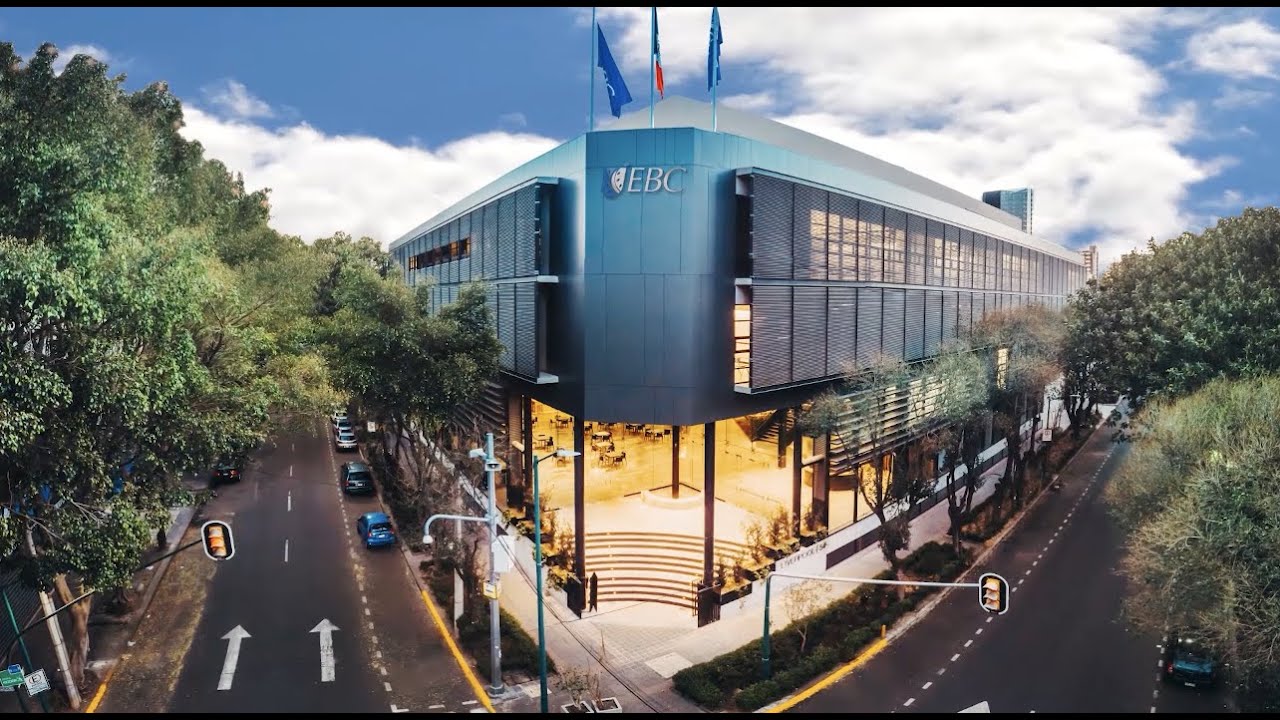 A modern building with the letters 'EBC' on the facade, surrounded by trees and situated at the intersection of two urban streets under a partly cloudy sky.