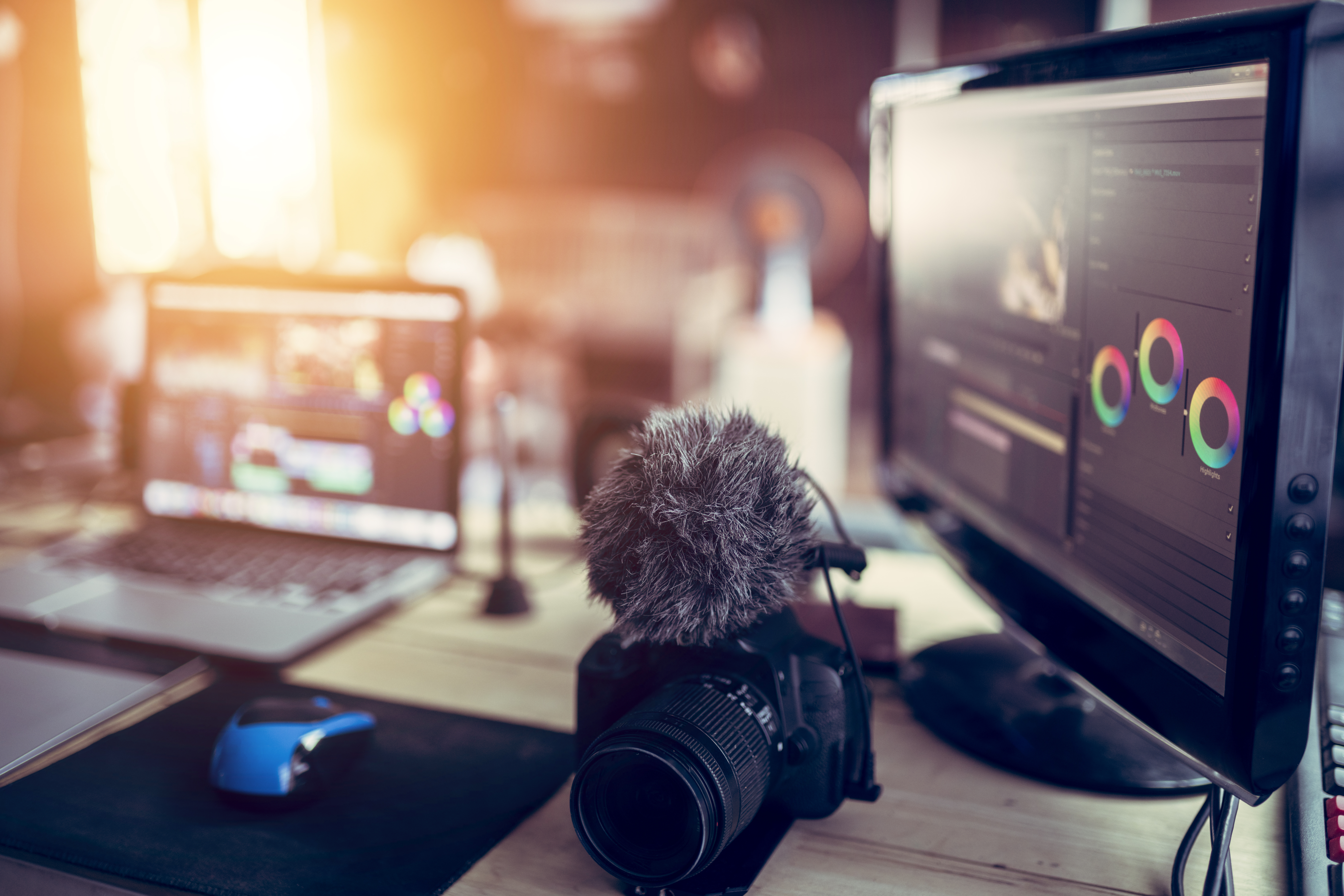 A workspace set up for freelance video editing, featuring a professional camera with an external microphone, a laptop displaying video editing software, a monitor with color grading tools, and a computer mouse on a desk, illuminated by natural lighting.