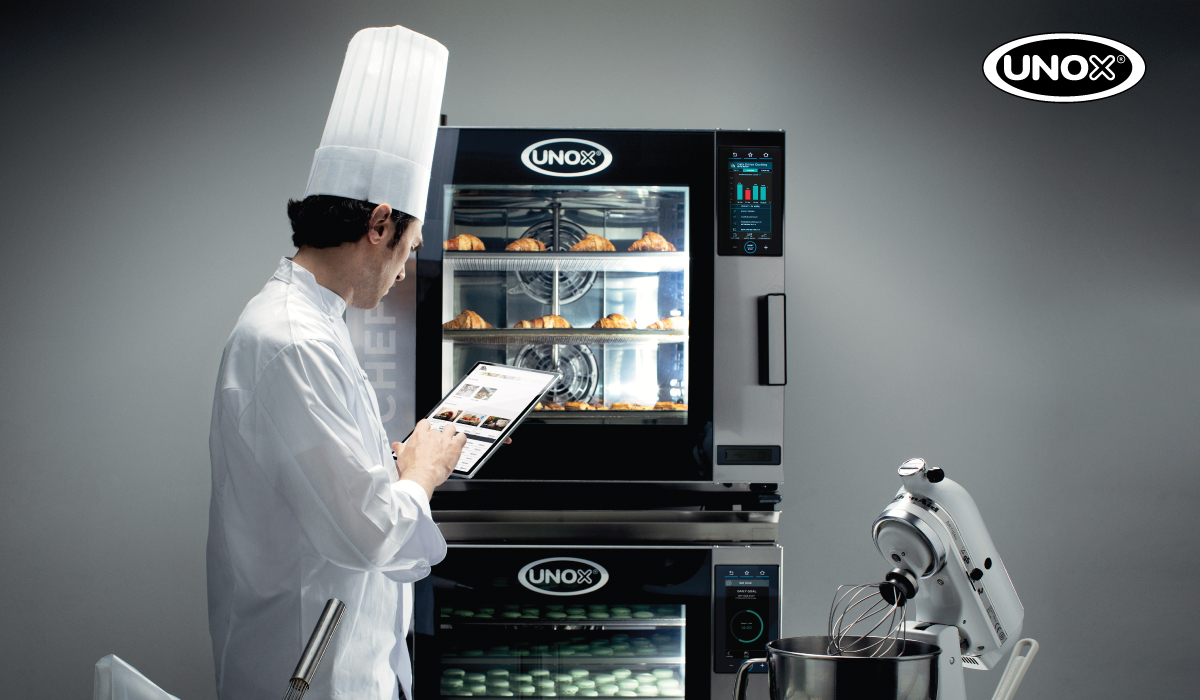 A chef in a white uniform and hat uses a digital tablet in front of a UNOX smart oven filled with baked goods. The modern kitchen setup includes another UNOX appliance and a stand mixer, highlighting the use of advanced kitchen technology and digital solutions in professional baking environments.