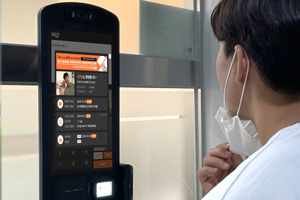 A person wearing a mask stands in front of a BROJ facial recognition access terminal, interacting with the device for access authentication.