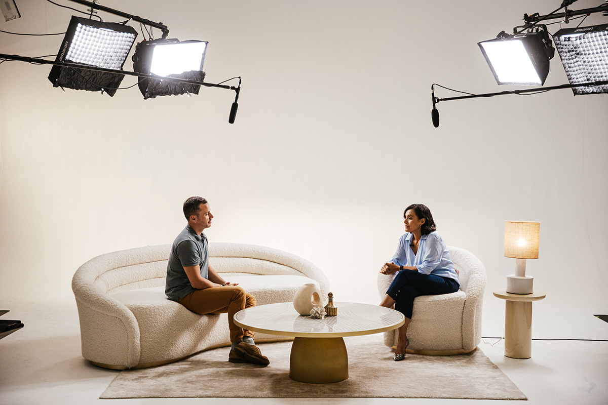 Two people seated on a modern white sofa set in a studio with bright lighting, a round coffee table, and a lamp on the side.