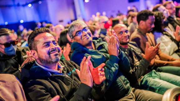 A group of attendees sits in an audience, clapping and enjoying a presentation at an AWS event. The scene is lively with people engaged and the atmosphere is positive.