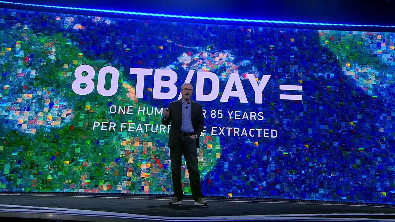 A keynote speaker stands on stage in front of a large display reading '80 TB/DAY = ONE HUMAN OVER 85 YEARS PER FEATURE EXTRACTED' during an AWS event. The screen features a colorful data mosaic background, emphasizing data processing capabilities.