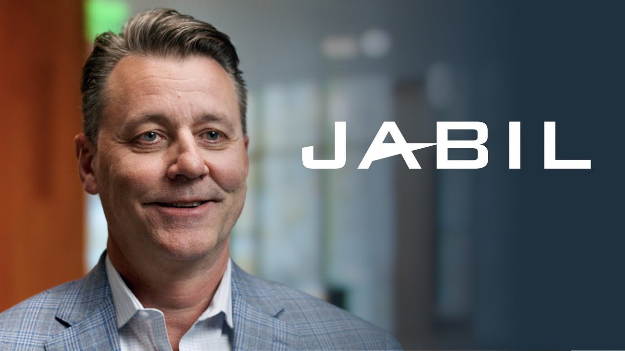 The word "Jabil" displayed in white text on a dark blue background, with a blurred office setting in the background.
