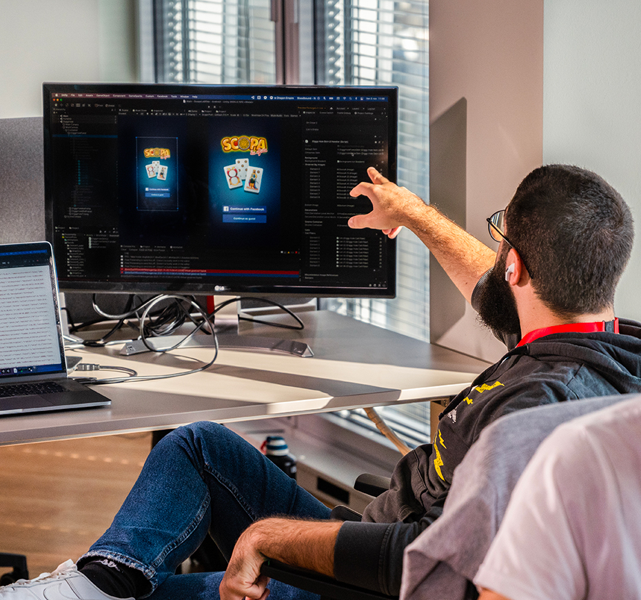 A developer collaborates with a colleague, pointing at a screen displaying a card game design in an office environment at Whatwapp. The scene highlights teamwork and digital game development.
