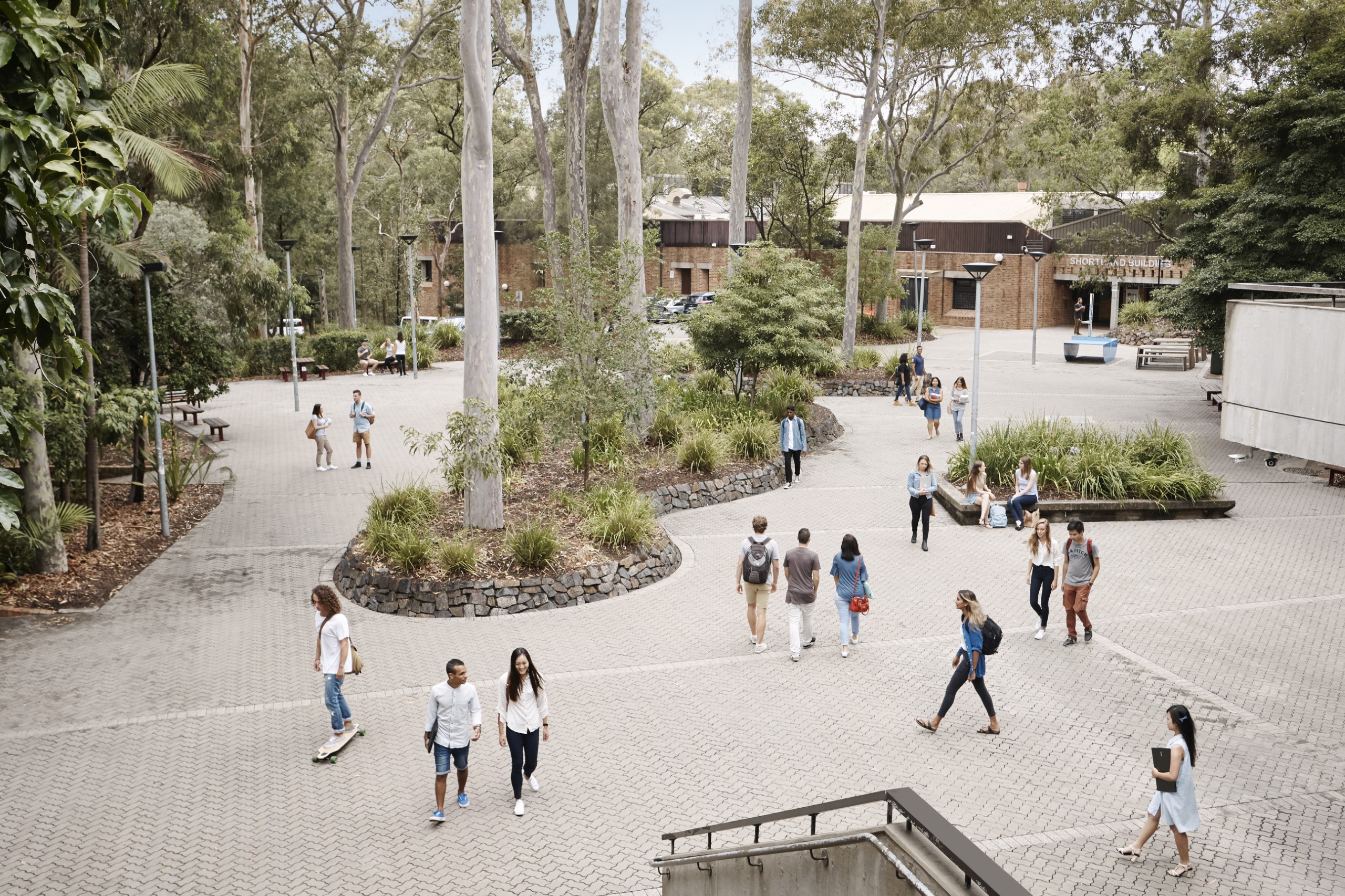 A university campus outdoor area with students walking and sitting among trees and landscaped gardens.