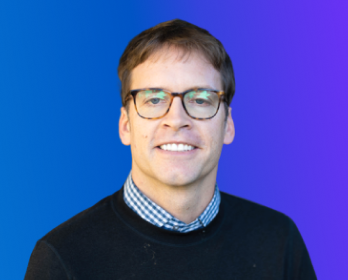 A man wearing glasses, a dark sweater, and a checkered shirt is smiling in front of a blue and purple gradient background.