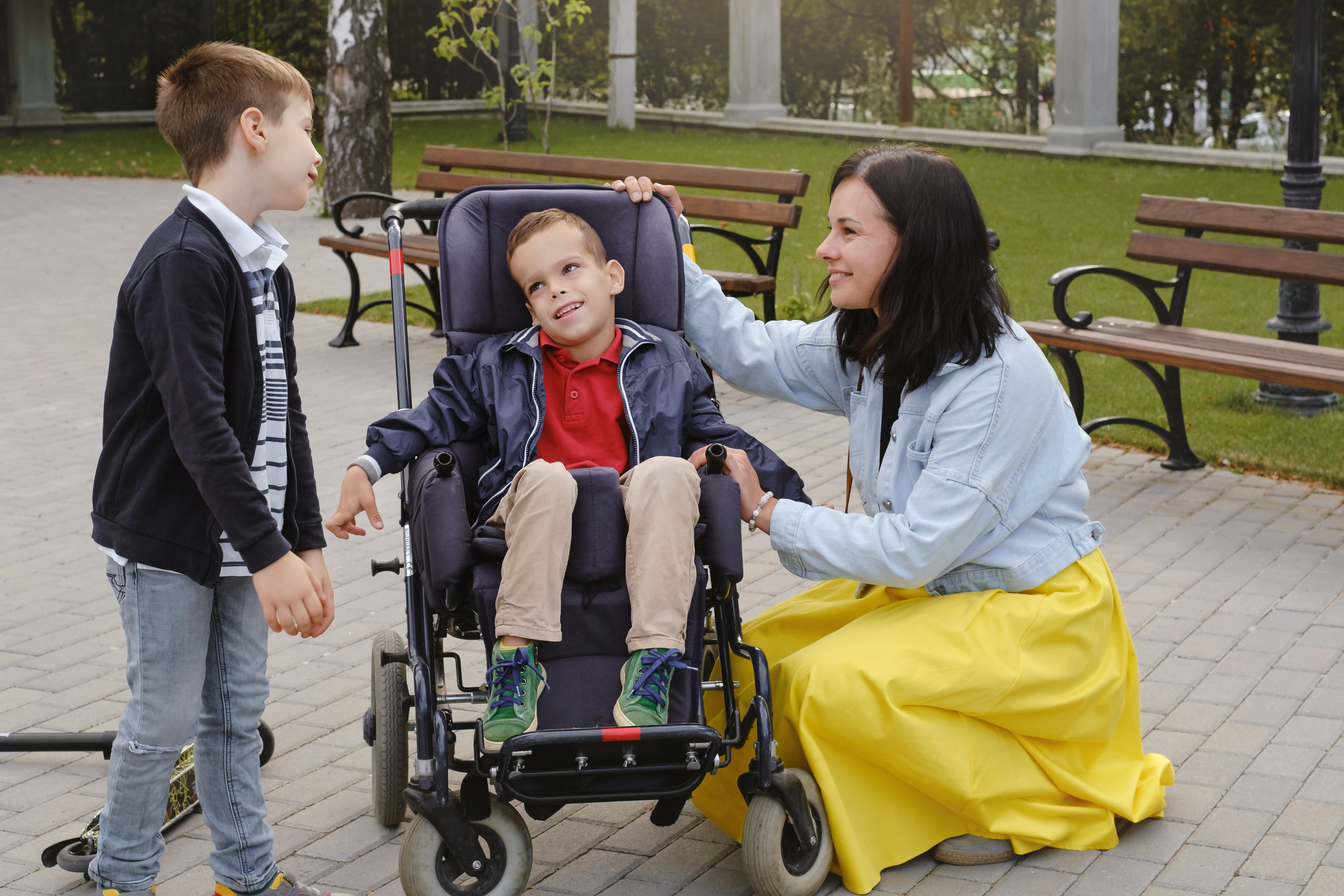 Family with cerebral palsy child on special wheelchair walking outdoors. Integration and accessibility of disabled people, inclusion. Brother with physical disorder Mother taking care of handicap kid