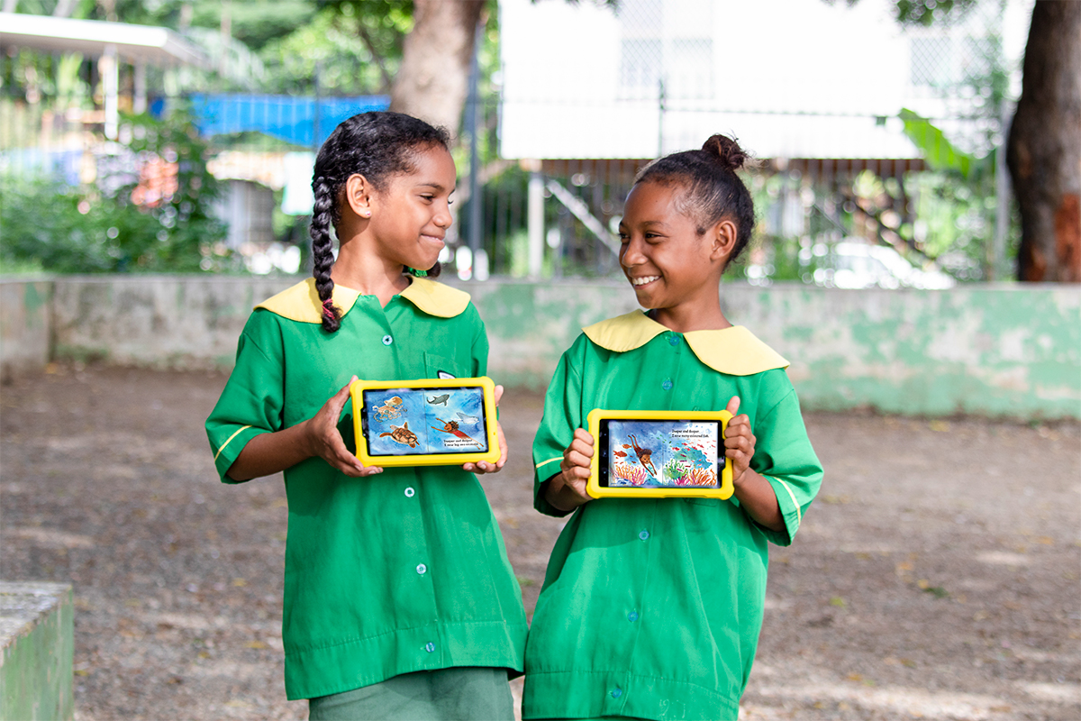 Two students in green uniforms are standing outdoors, smiling at each other while holding tablets with educational content on the screens.