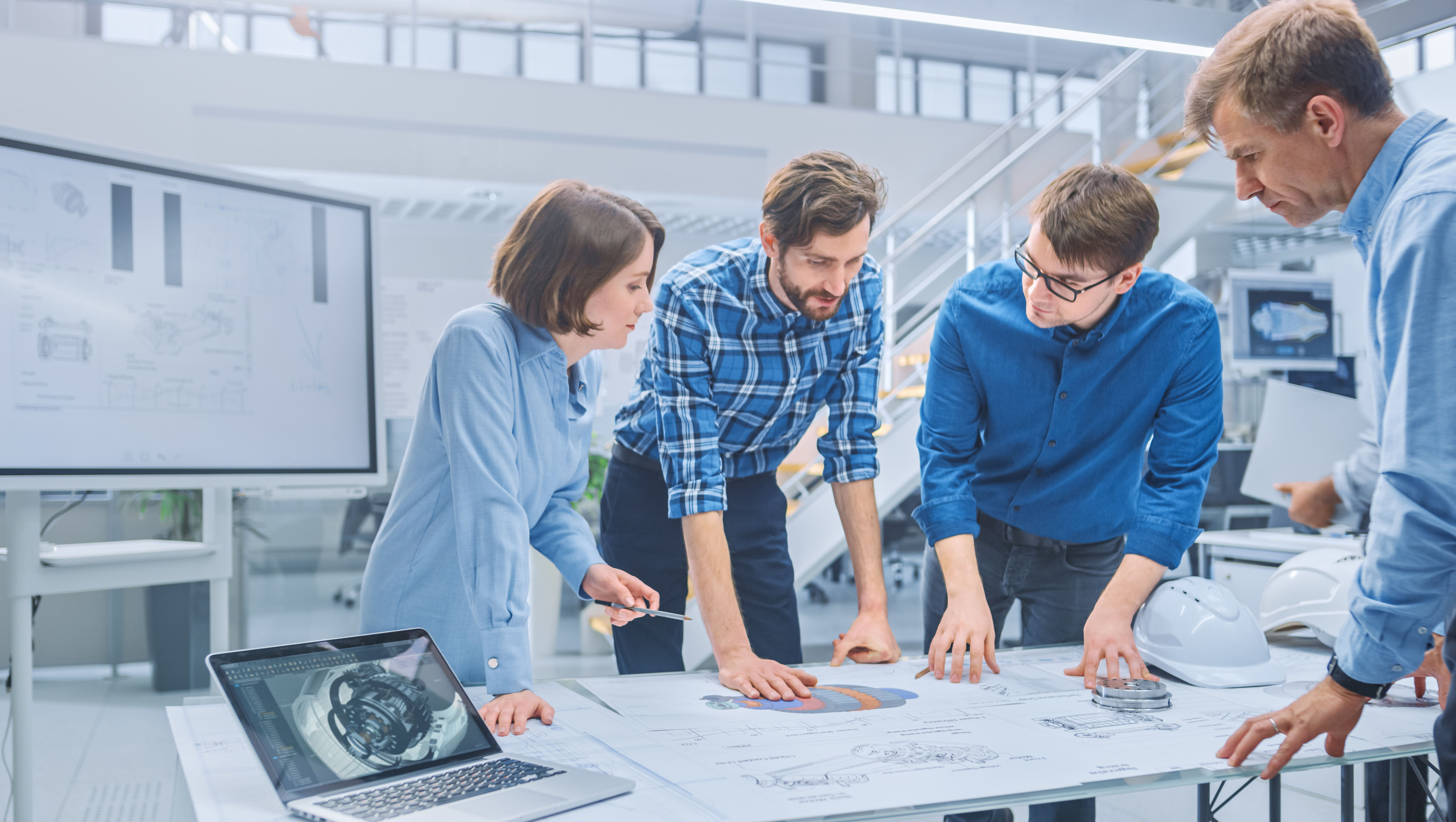 A group of engineers collaborating on a project around a large table with technical drawings, a laptop displaying a 3D model, and engineering diagrams on a screen in a modern office environment.