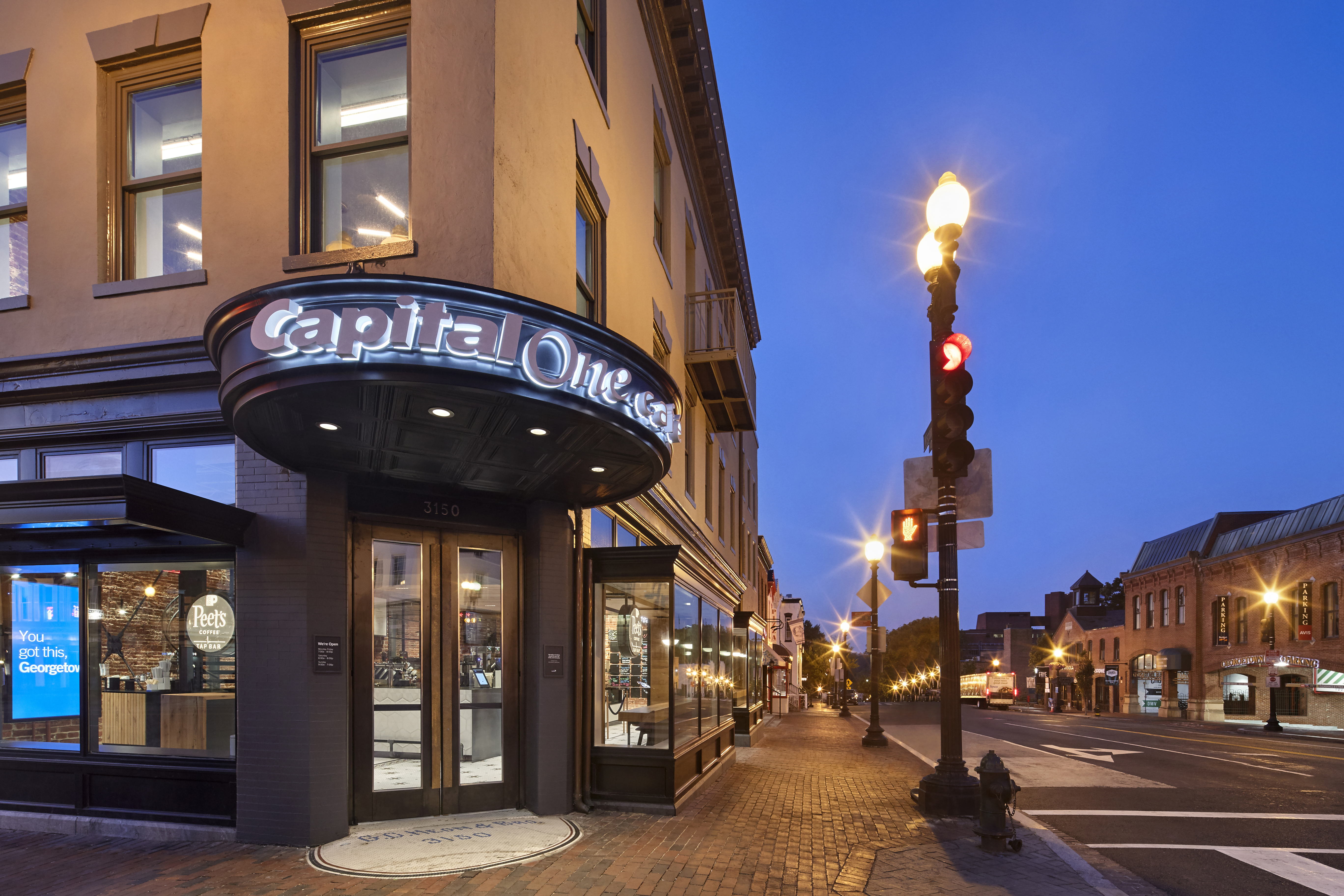 Exterior view of a Capital One Café in Georgetown at night, with illuminated signage, large windows, and a city street scene including a stoplight and brick buildings.