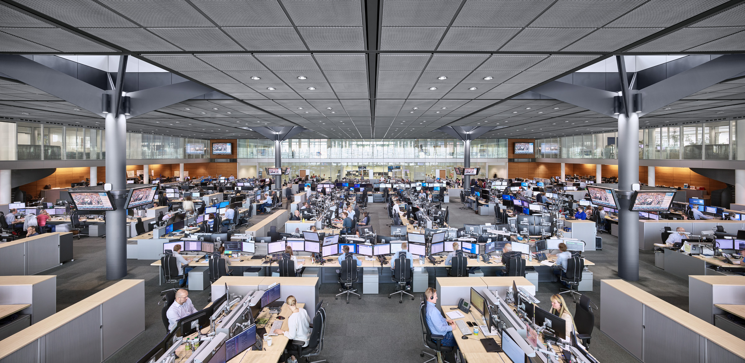 A spacious, modern open-plan office with numerous employees working at grouped desks filled with computer monitors and office equipment. Large windows and high ceilings provide a bright, collaborative workspace environment.