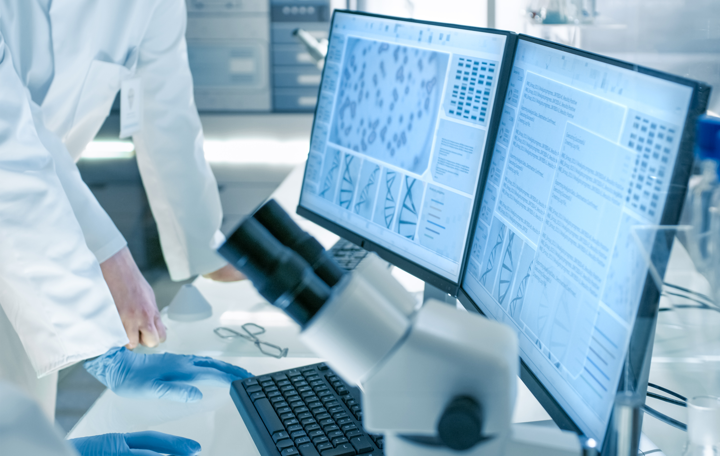 Laboratory researchers in lab coats and gloves analyze genetic and microscopic data displayed on dual computer monitors, with a microscope in the foreground.