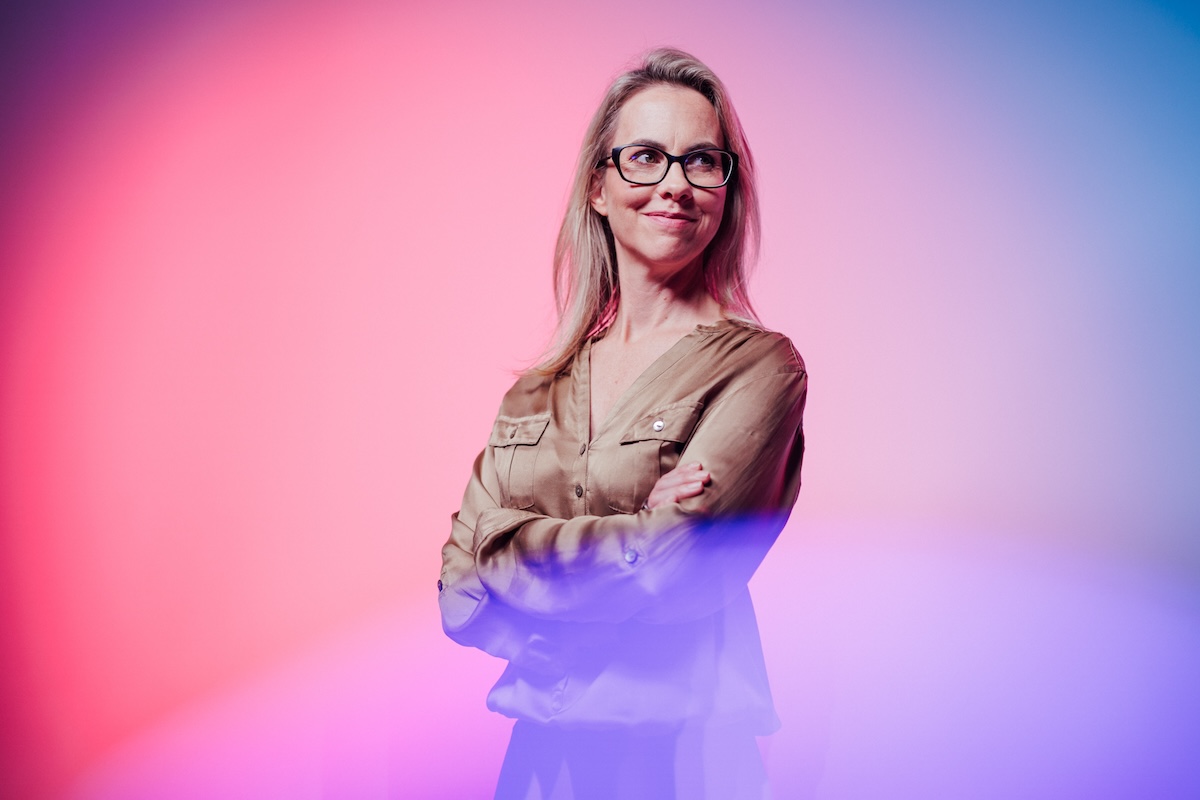 A confident woman wearing glasses stands with arms crossed in front of a vibrant, colorful gradient background.