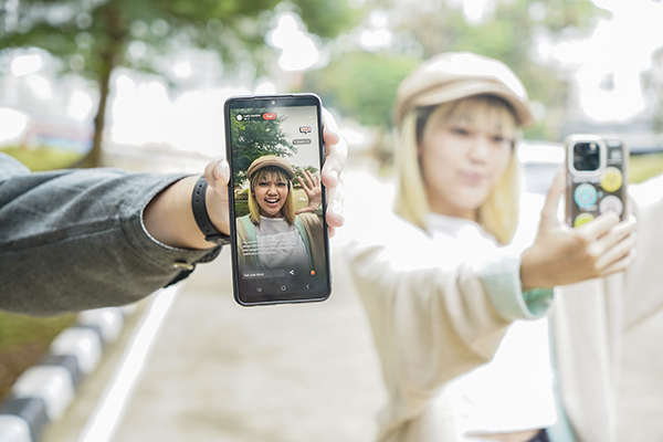A person holding a smartphone displaying a live video stream outdoors, with another person taking a selfie in the background.