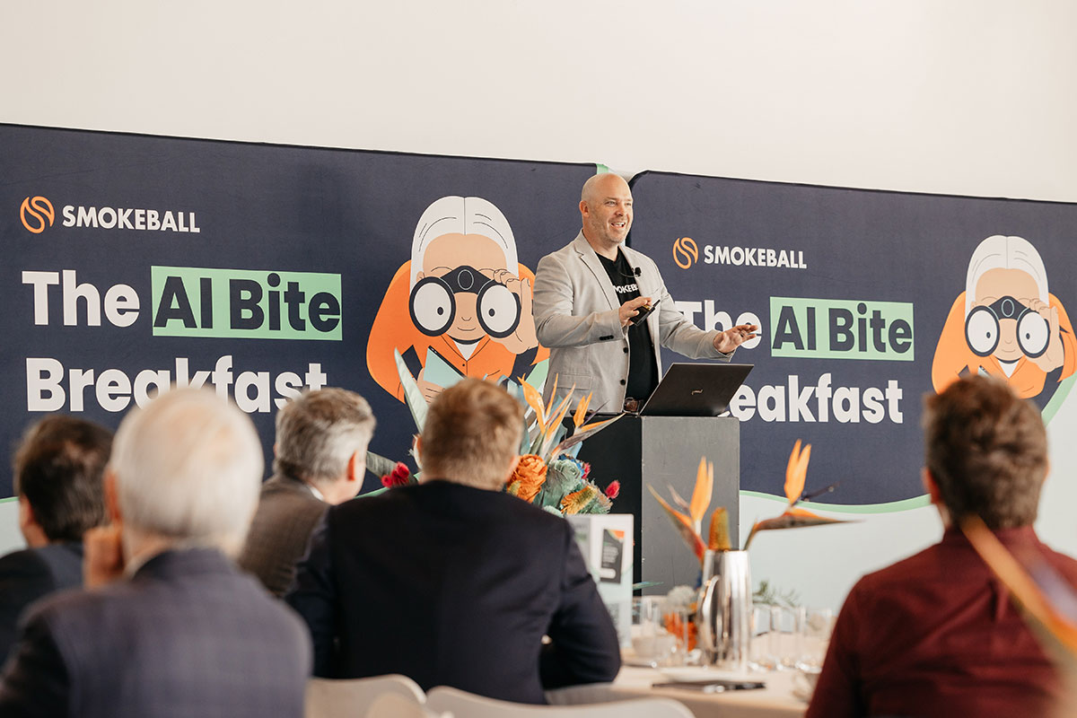 A speaker presenting at "The AI Bite Breakfast" event, with a colorful banner and audience seated at tables with floral centerpieces.