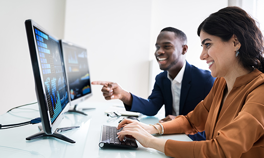 Two business professionals analyzing data and graphs on computer monitors in an office environment.