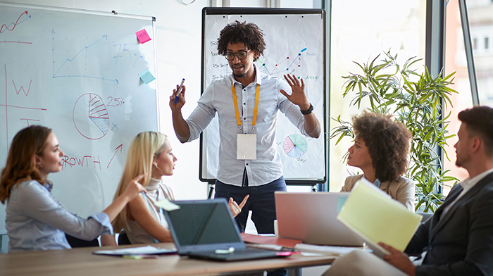 A group of people in a modern office setting engaged in a presentation, with charts and graphs displayed on a whiteboard and flipchart.