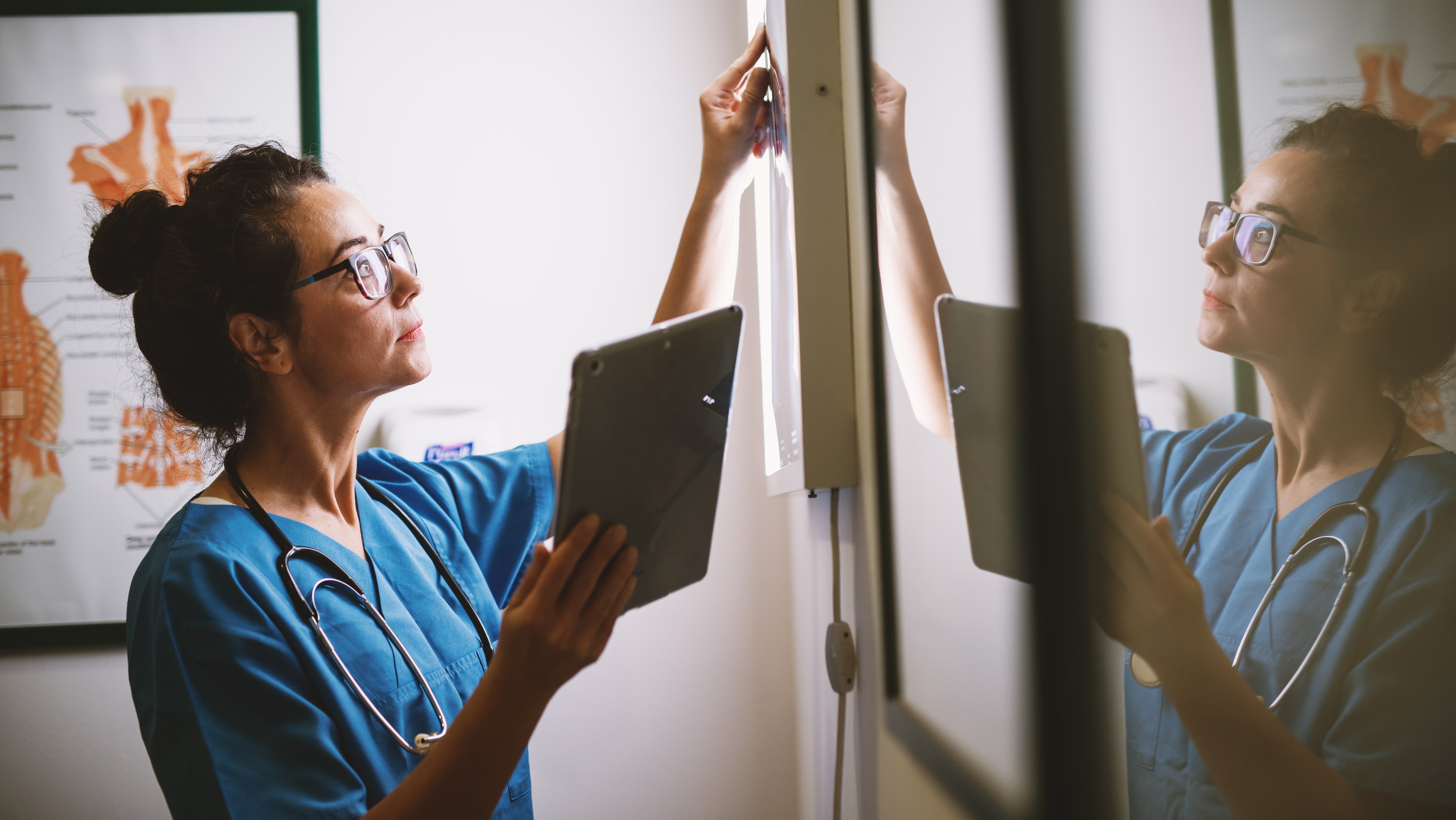 A healthcare professional in scrubs with a stethoscope examines a medical scan on a lightbox while holding a tablet, in a clinical setting.