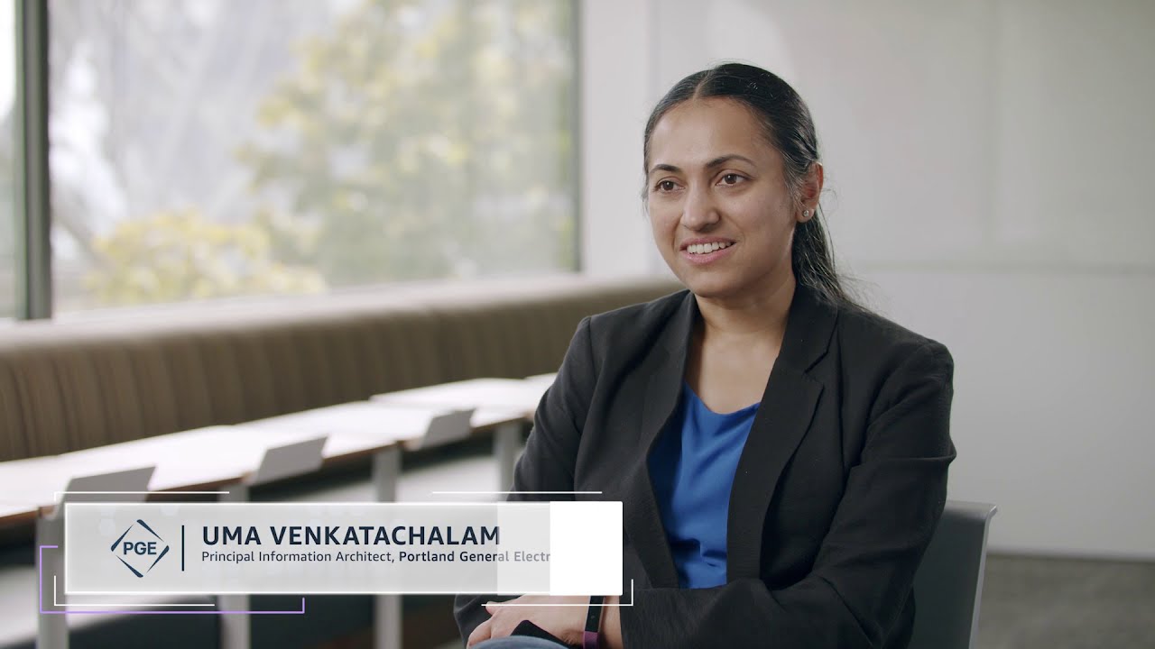 A professional woman in a business suit is seated and speaking in an interview setting. The background shows a modern office environment with large windows and natural light. A text overlay indicates she is the Principal Information Architect at Portland General Electric.