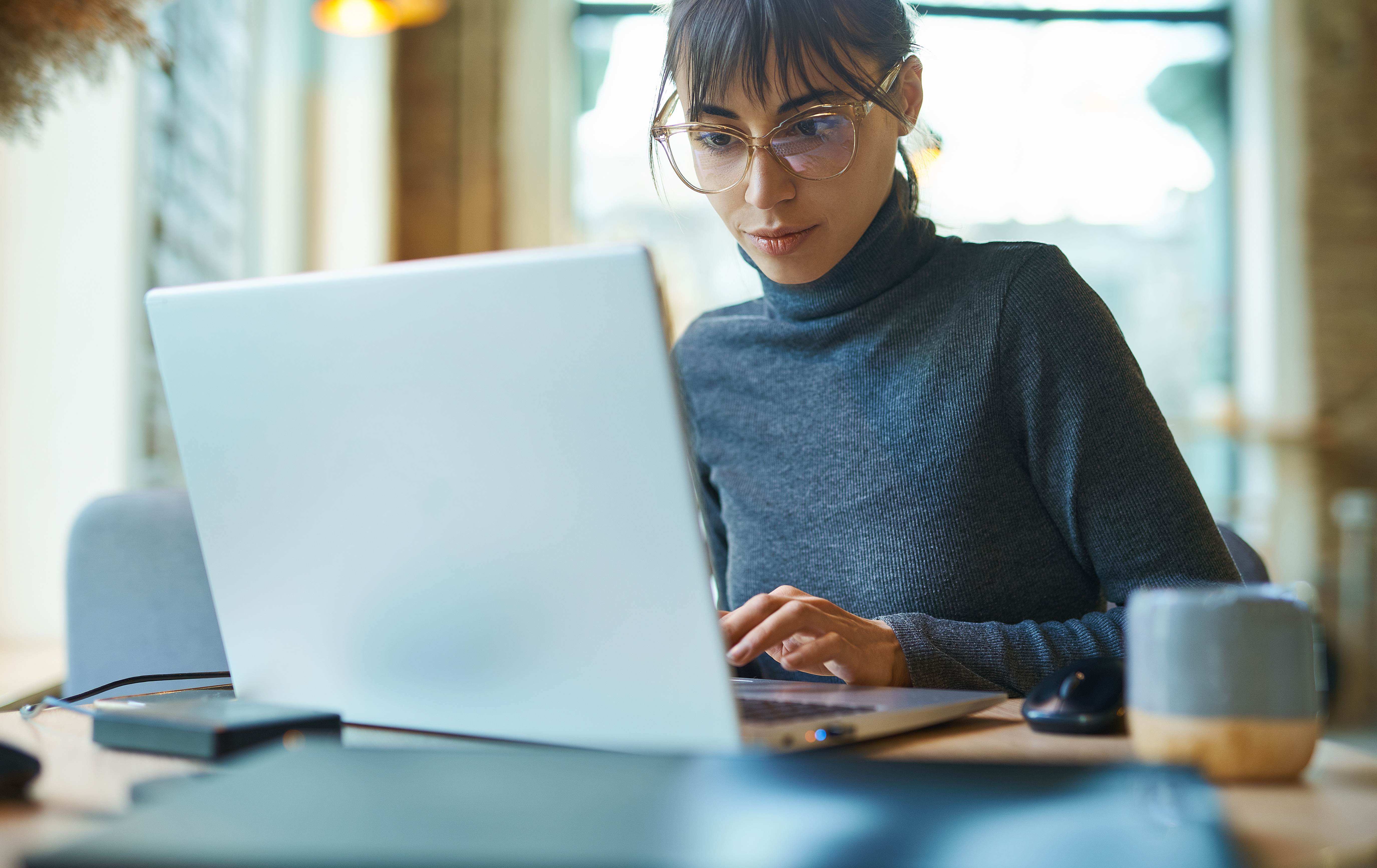 portrait of skilled young female developer in eyeglasses concentrating on screen and typing on laptop while sitting at desk at workplace or cafe. Concept remote work, freelance, using laptop computer.