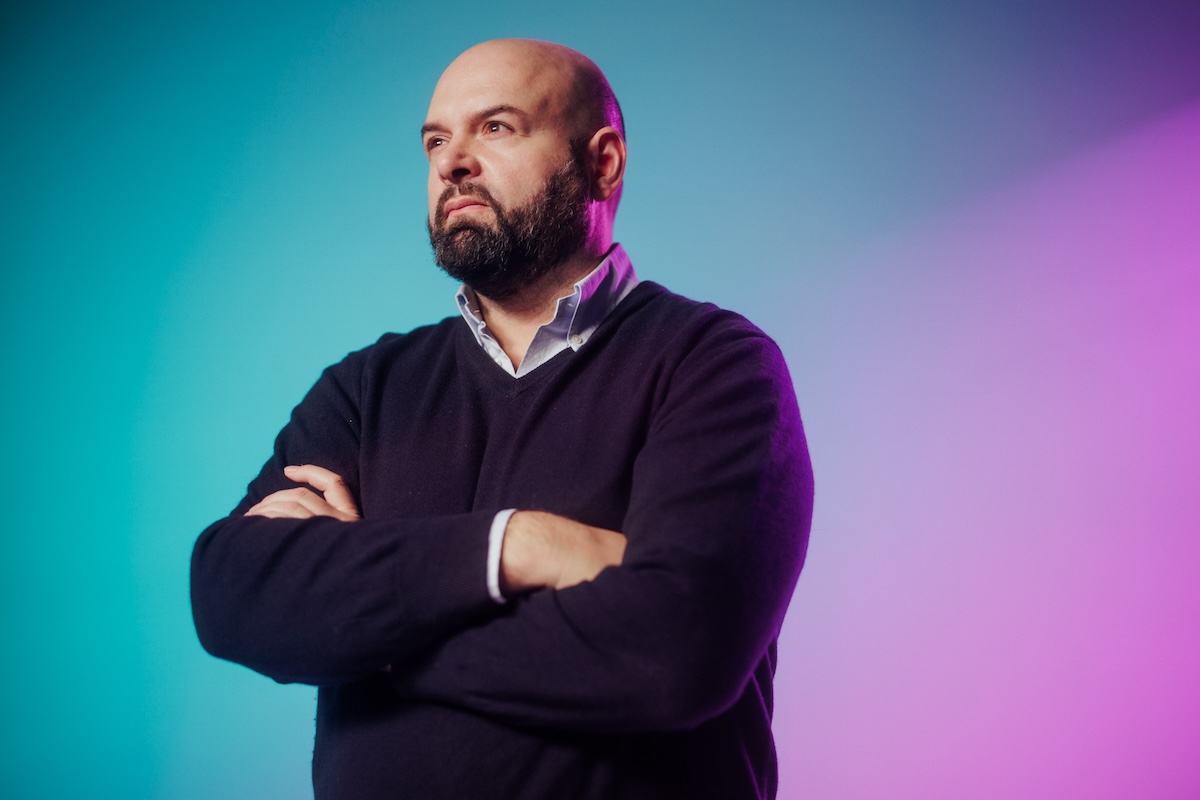 A bald man with a beard, wearing a dark sweater and collared shirt, stands with his arms crossed against a gradient blue and purple background.