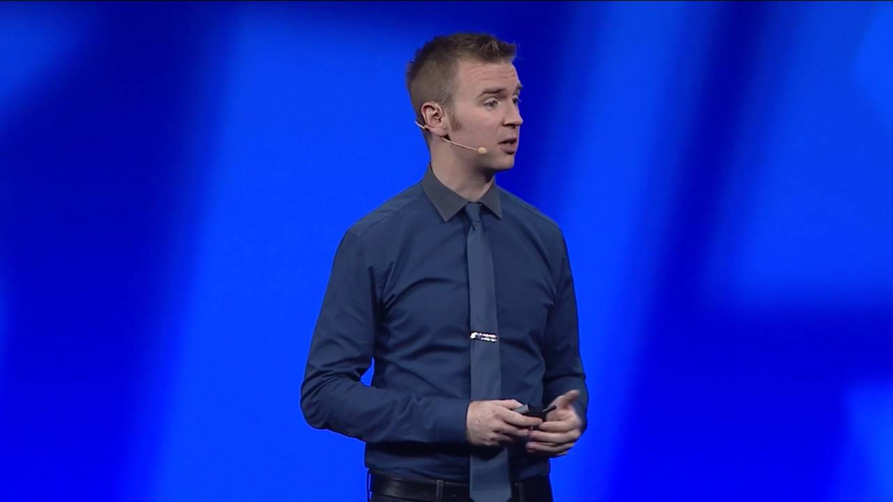 A man wearing a blue shirt and tie giving a presentation on stage with a blue background.