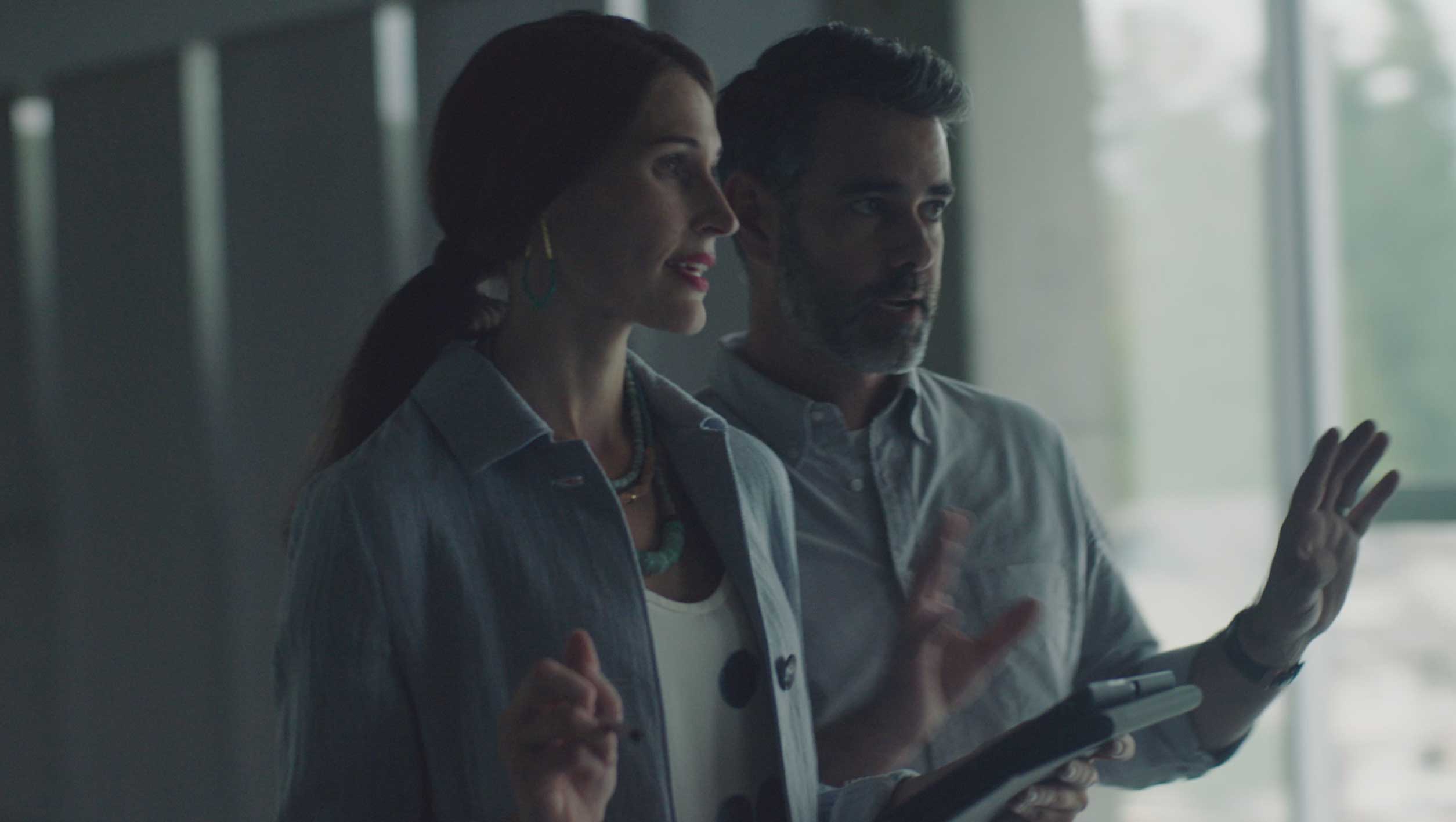 Two professionals standing and talking inside a modern office, with one holding a tablet device. The scene is softly lit with large windows in the background.