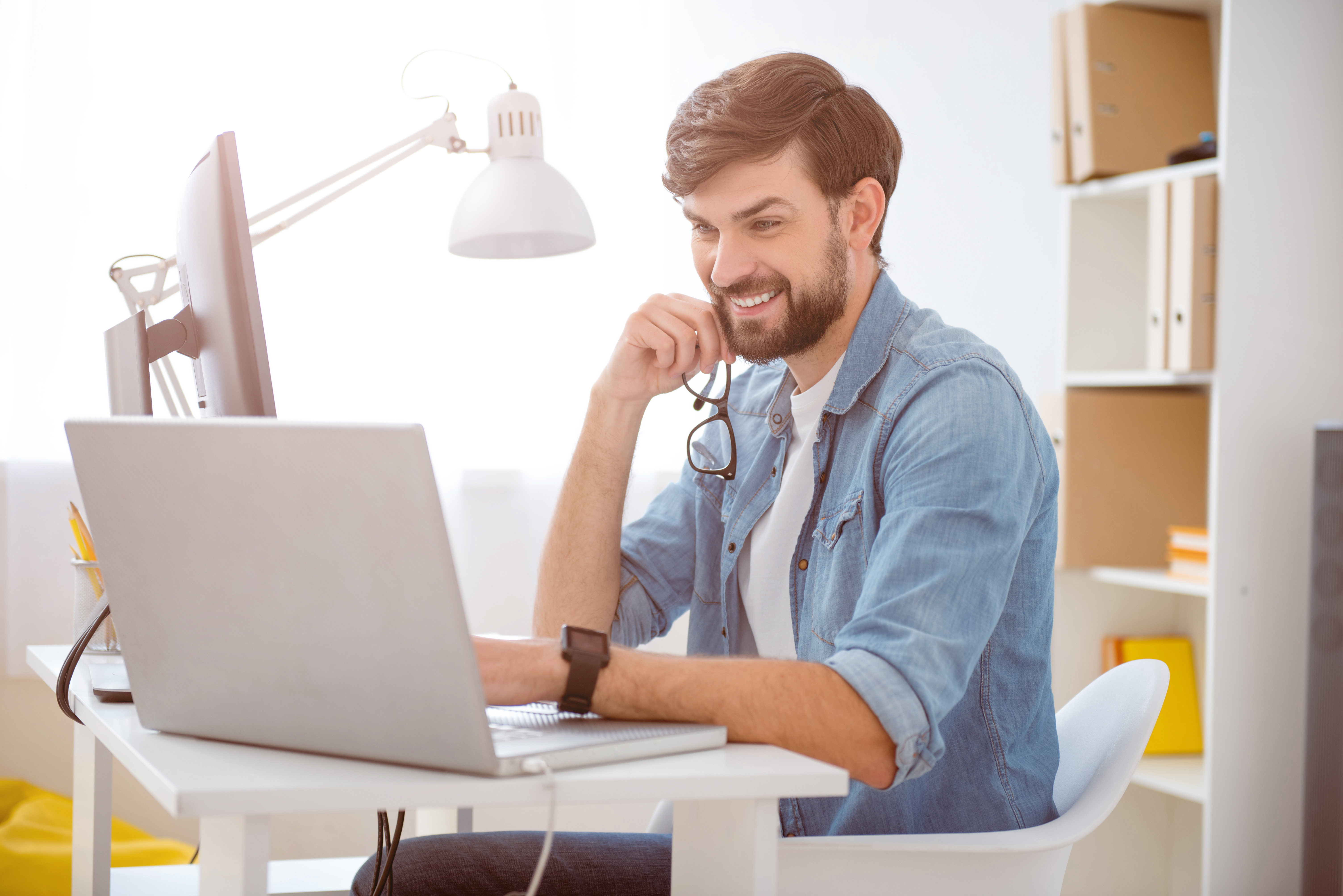 In a good mood. Young bearded guy smiling while using his laptop and holding glasses in his hand