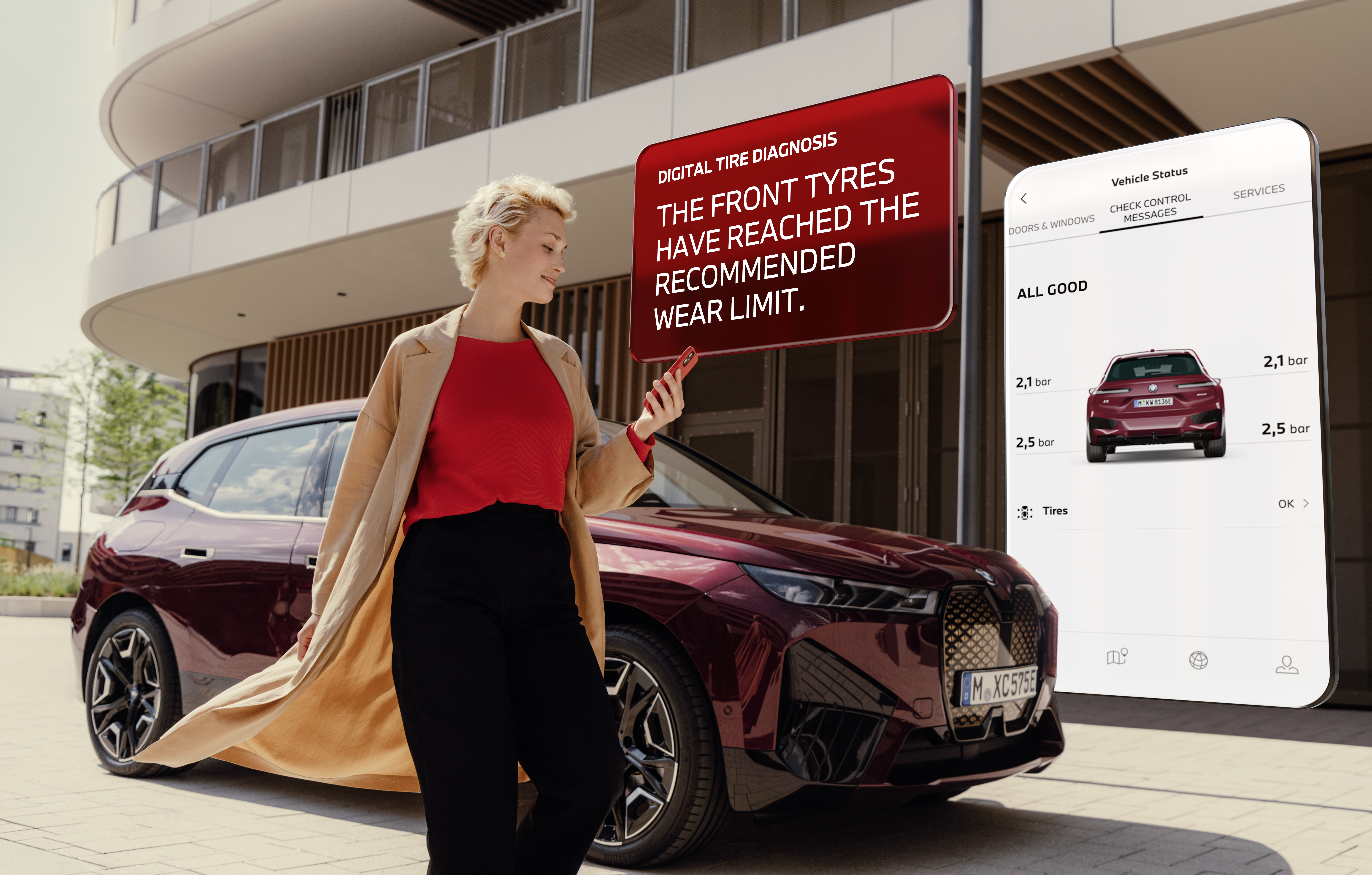 A woman stands beside a maroon car, holding a smartphone that displays a digital tire diagnosis alert: 'The front tyres have reached the recommended wear limit.' A visual overlay shows a mobile app with vehicle status and tire information. Modern building in the background, showcasing connected automotive technology.