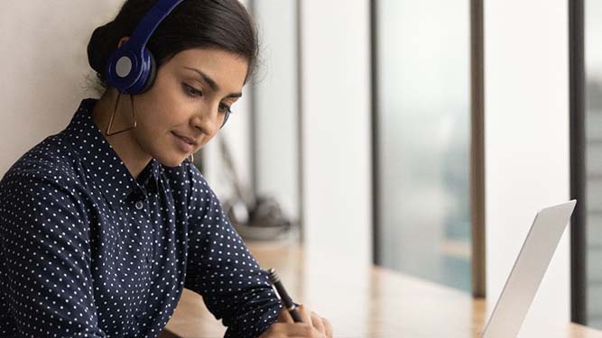 A student wearing headphones sits at a desk, writing in a notebook while using a laptop for remote learning. Large windows are in the background, suggesting a modern learning environment.