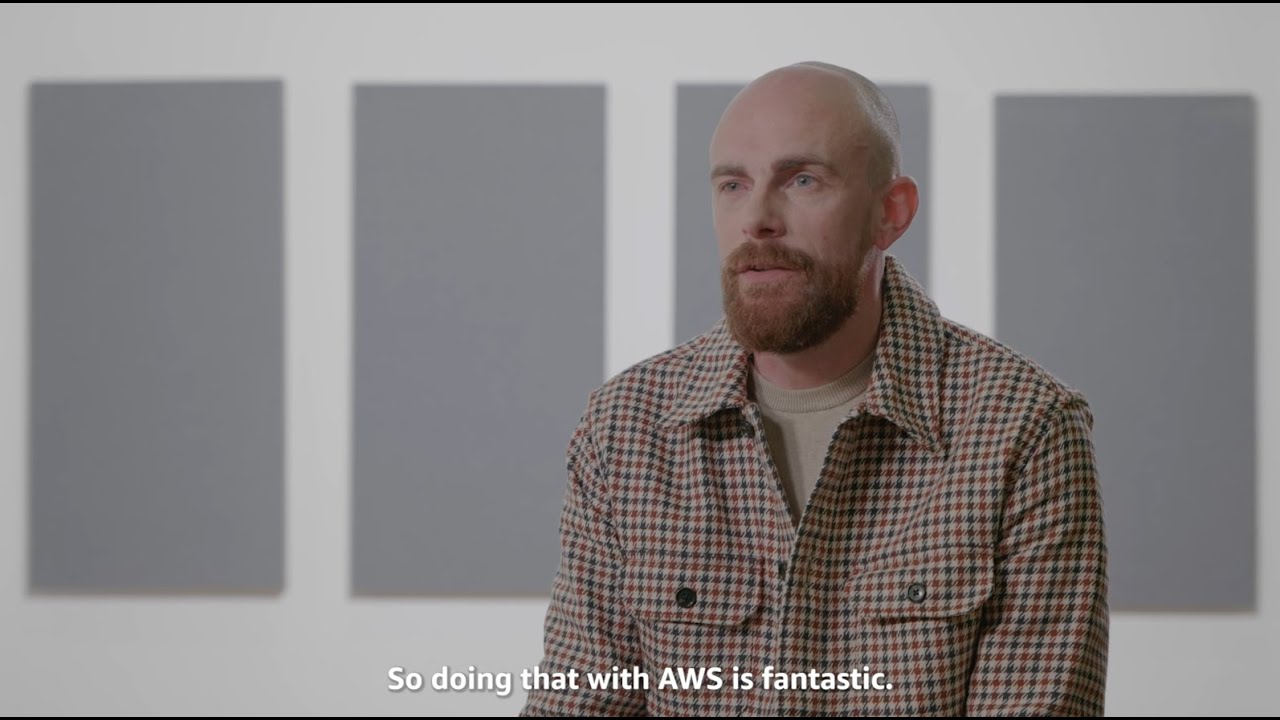 A man is interviewed in front of an abstract background, with on-screen text reading 'So doing that with AWS is fantastic.'