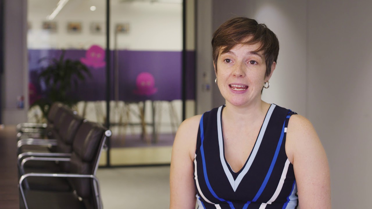 A person speaking in a modern conference room or office, with glass walls, chairs, and a blurred meeting area in the background.