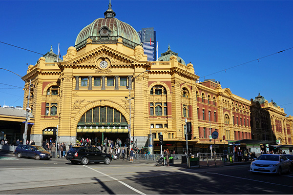 Flinders Street Station in Melbourne, Australia, a historic railway station with distinctive yellow facade and green dome, busy with people and traffic in front.