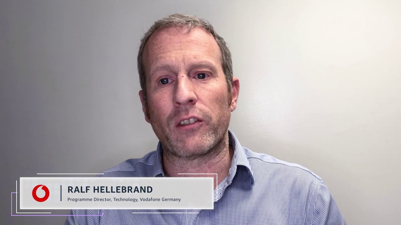 A man in a business shirt is speaking in front of a neutral background. On the lower left, a title box displays the Vodafone logo and the text 'RALF HELLEBRAND, Programme Director, Technology, Vodafone Germany.'