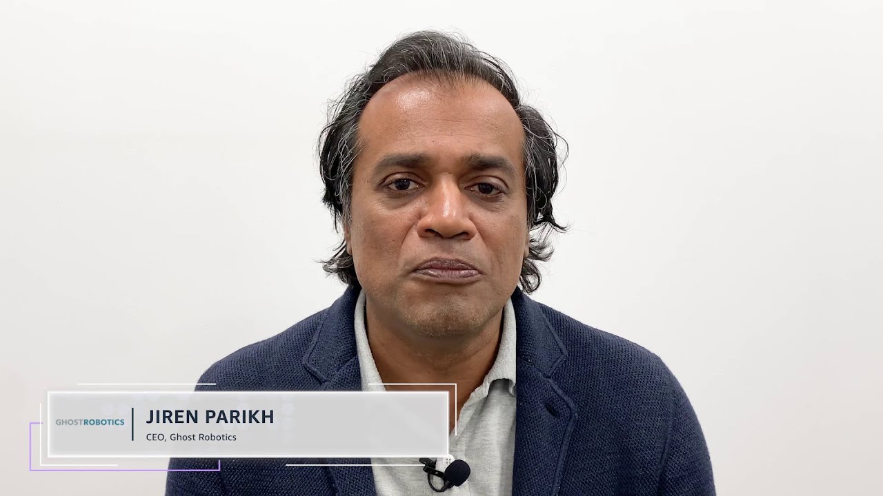 Jiren Parikh, CEO of Ghost Robotics, is seated for an interview or presentation, wearing a navy blazer and a light shirt, with a microphone clipped to his collar against a white background.