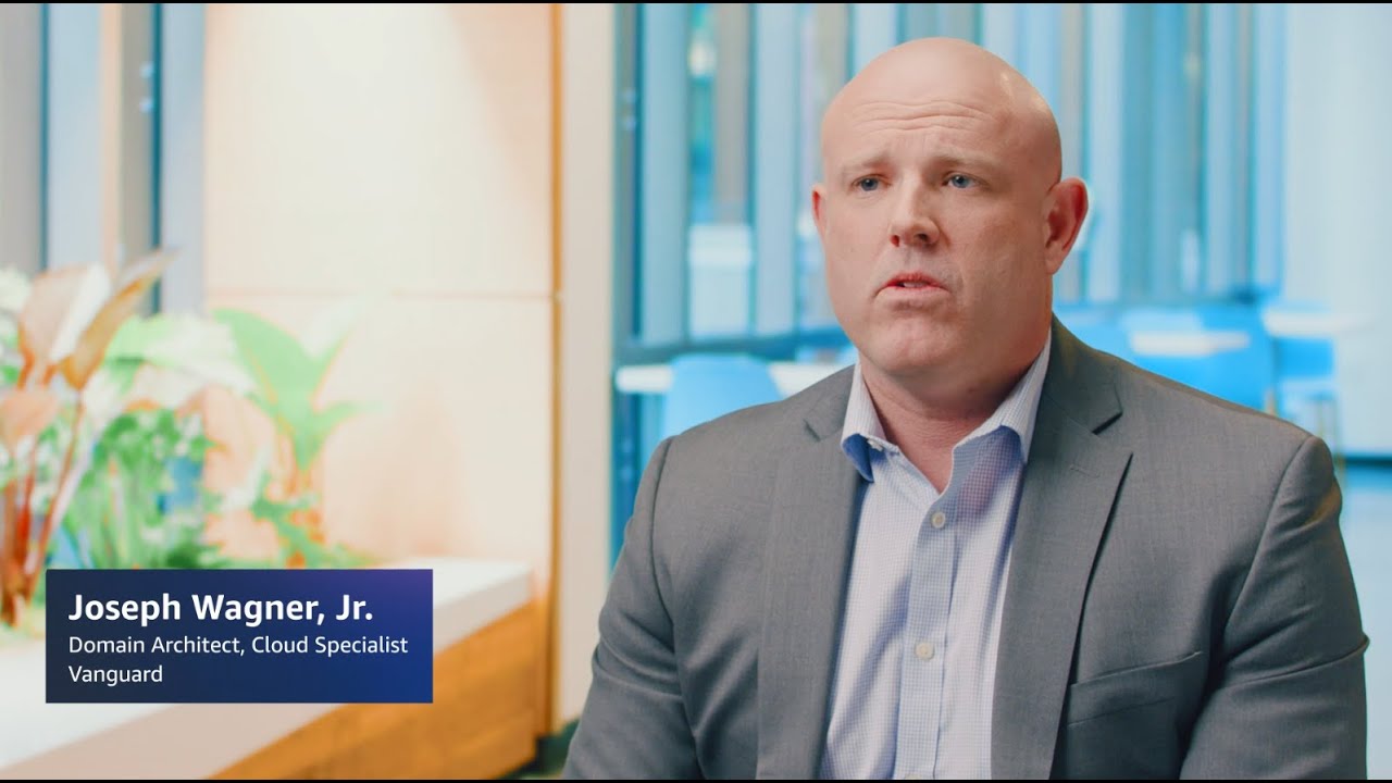 A professional man in a suit, seated for an interview, with text on the image saying 'Joseph Wagner, Jr. - Domain Architect, Cloud Specialist, Vanguard.' There are plants and a blurred office background.