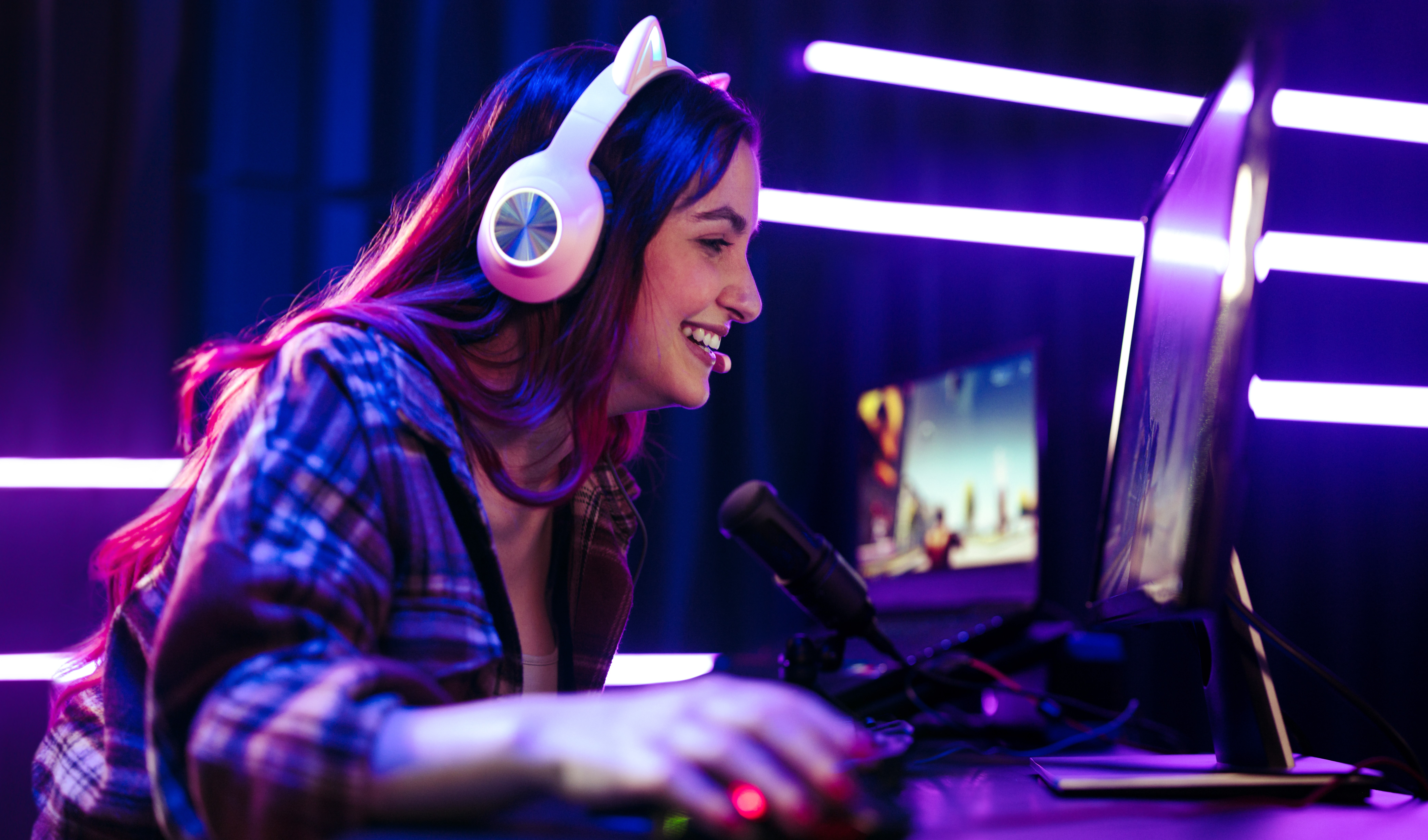 A young person wearing a headset with cat ears, smiling and playing a PC game in a neon-lit room with a microphone and monitor.