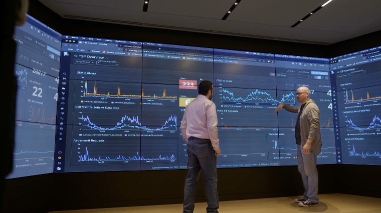 Two men stand in front of a large wall of screens displaying various data charts and graphs in a modern, tech-focused environment.