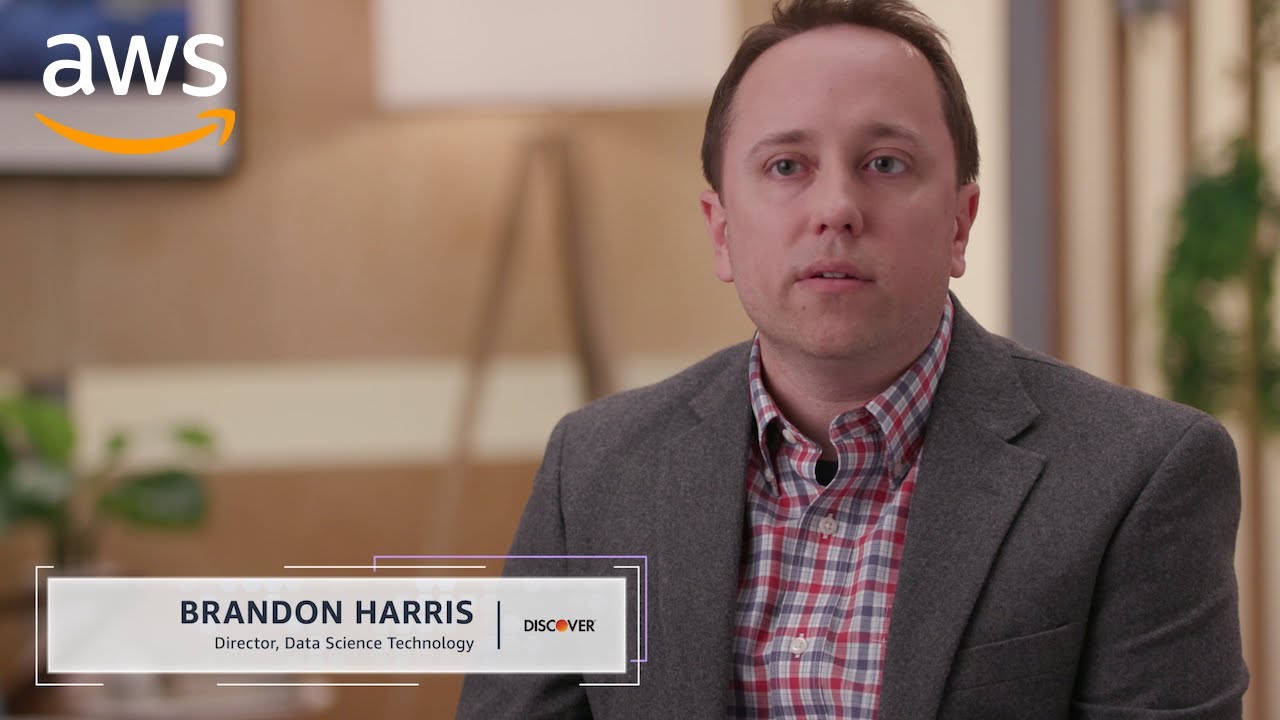 A professional interview setting featuring an individual identified as the Director of Data Science Technology at Discover, filmed for an AWS solution case study. The subject is seated, wearing a gray blazer and plaid shirt, with AWS branding visible in the corner.