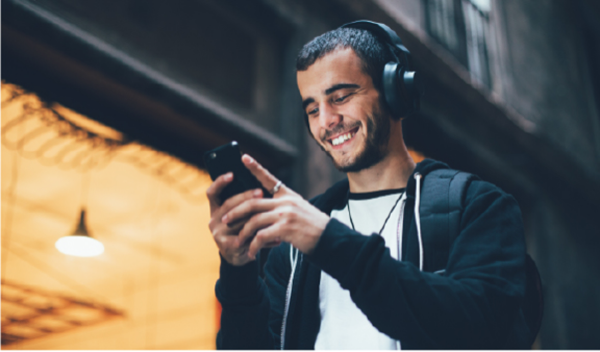 A young man wearing headphones smiles as he looks at his smartphone while standing outdoors in an urban environment.