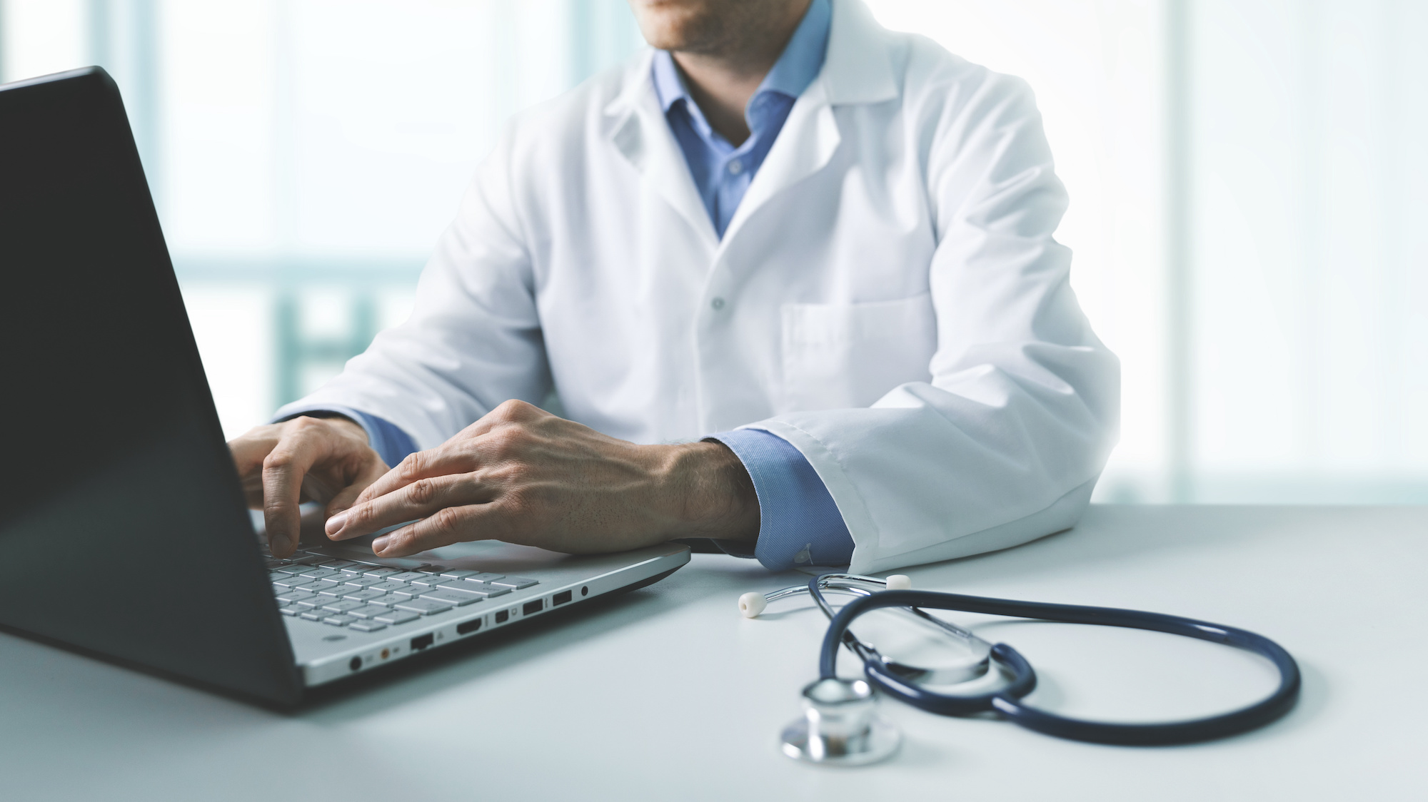 A doctor in a white lab coat working on a laptop at a desk with a stethoscope nearby, representing healthcare professionals using technology.