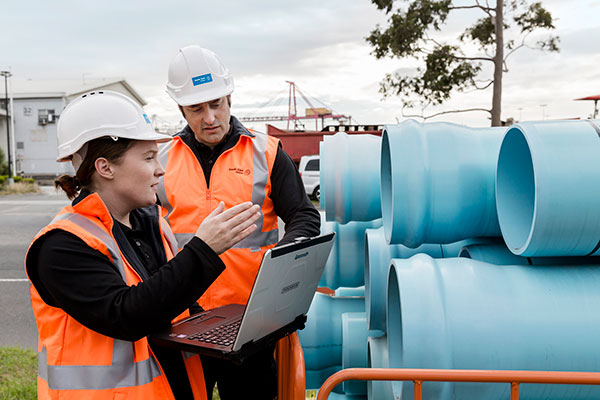 Two workers in safety gear inspecting large blue water pipes while using a laptop, representing AI-driven pressure anomaly inspection at South East Water.