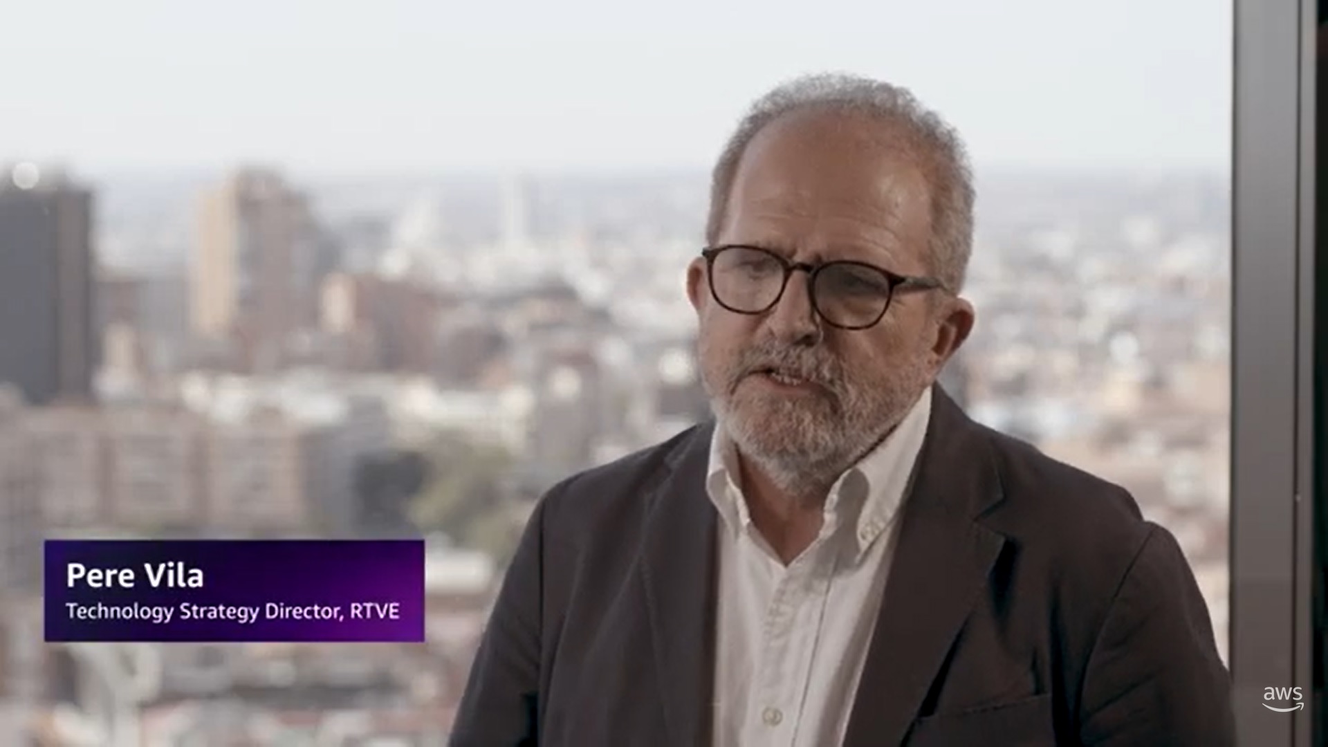 A person in a suit and glasses is interviewed in front of a large city window. The name 'Pere Vila' and title 'Technology Strategy Director, RTVE' are shown in a graphic overlay. The background is softly blurred urban cityscape.