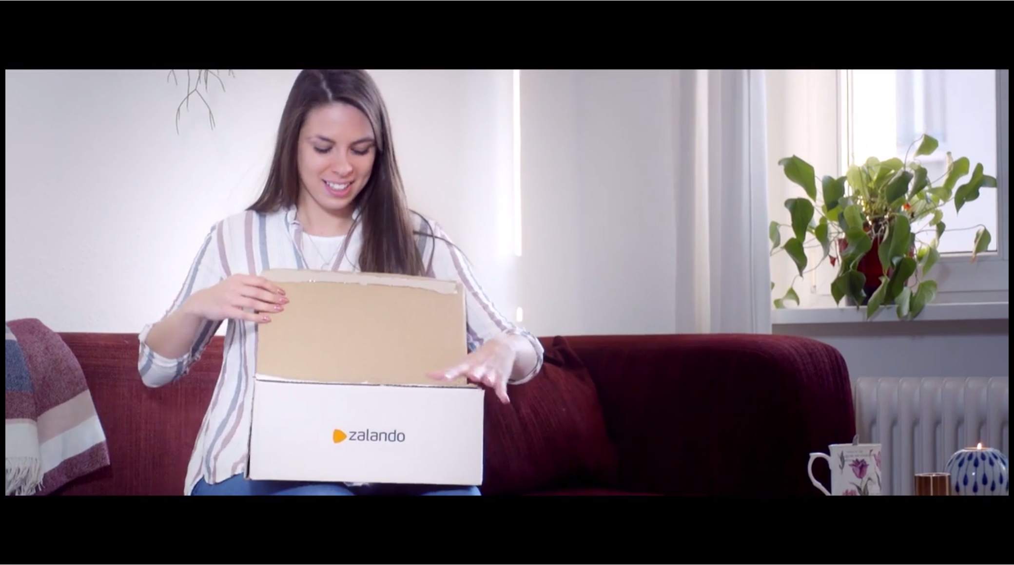 A woman sitting on a red sofa opens a Zalando cardboard box with a smile. The scene includes a blanket, indoor plant on a windowsill, and a teacup with a candle on a table nearby.