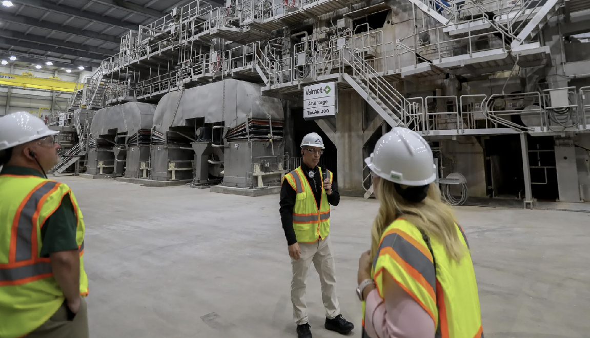 Workers wearing safety vests and hard hats participate in a tour of a Georgia-Pacific factory, standing in front of large industrial machinery inside a modern manufacturing facility.
