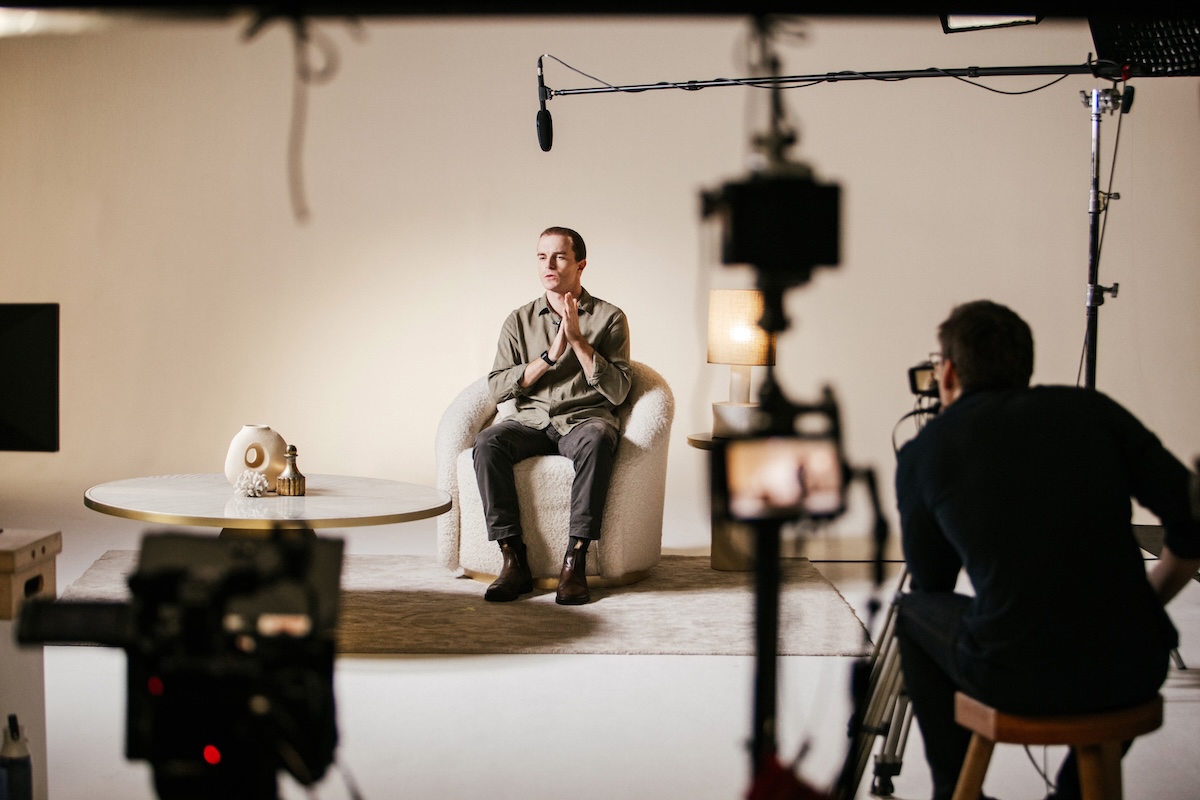 A behind-the-scenes look at a video interview setup, featuring a person seated in a chair under studio lighting, with cameras, a microphone, and a crew visible in the foreground.