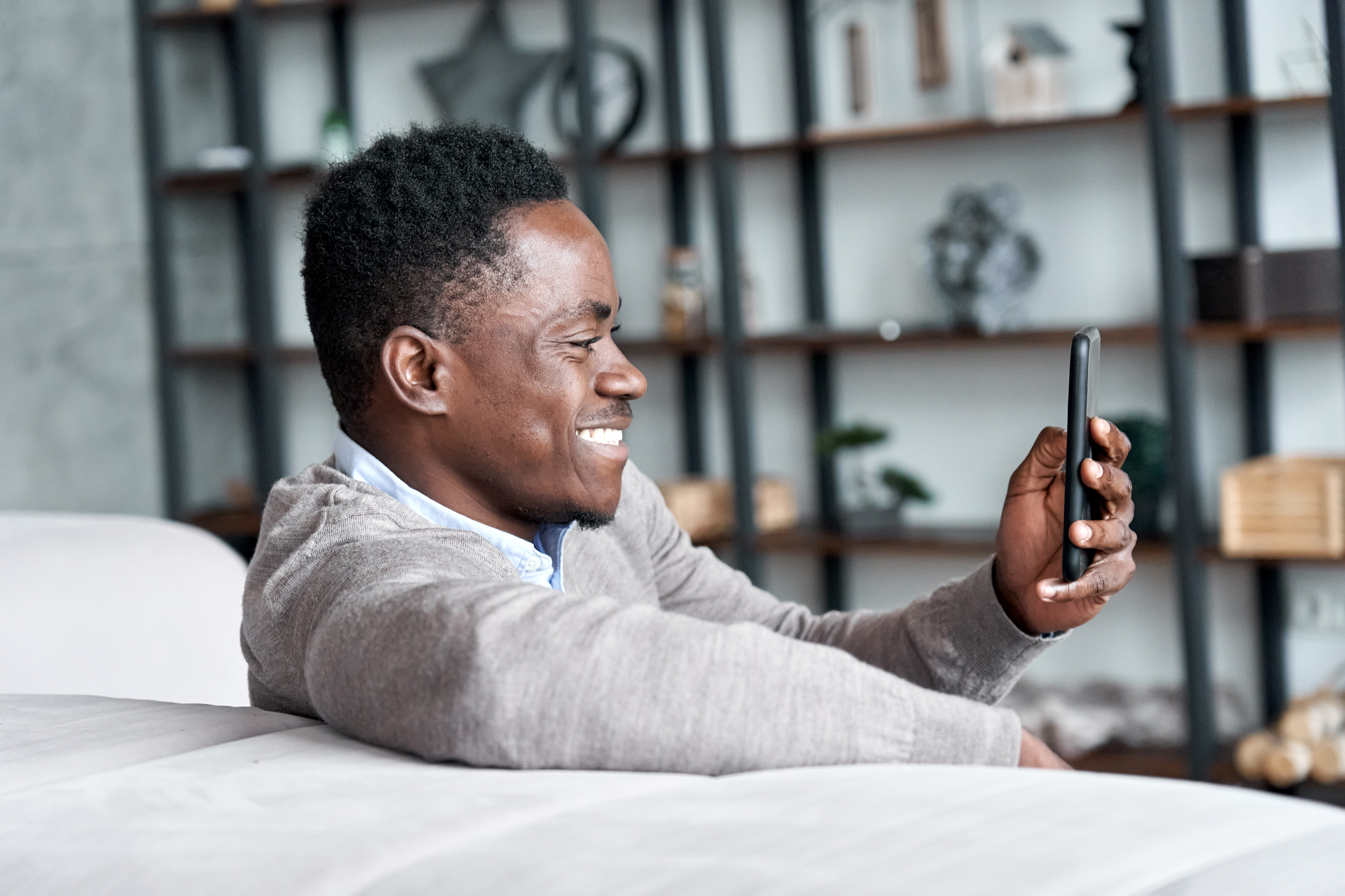 A man sitting on a couch, smiling and looking at his smartphone in a modern living room setting.
