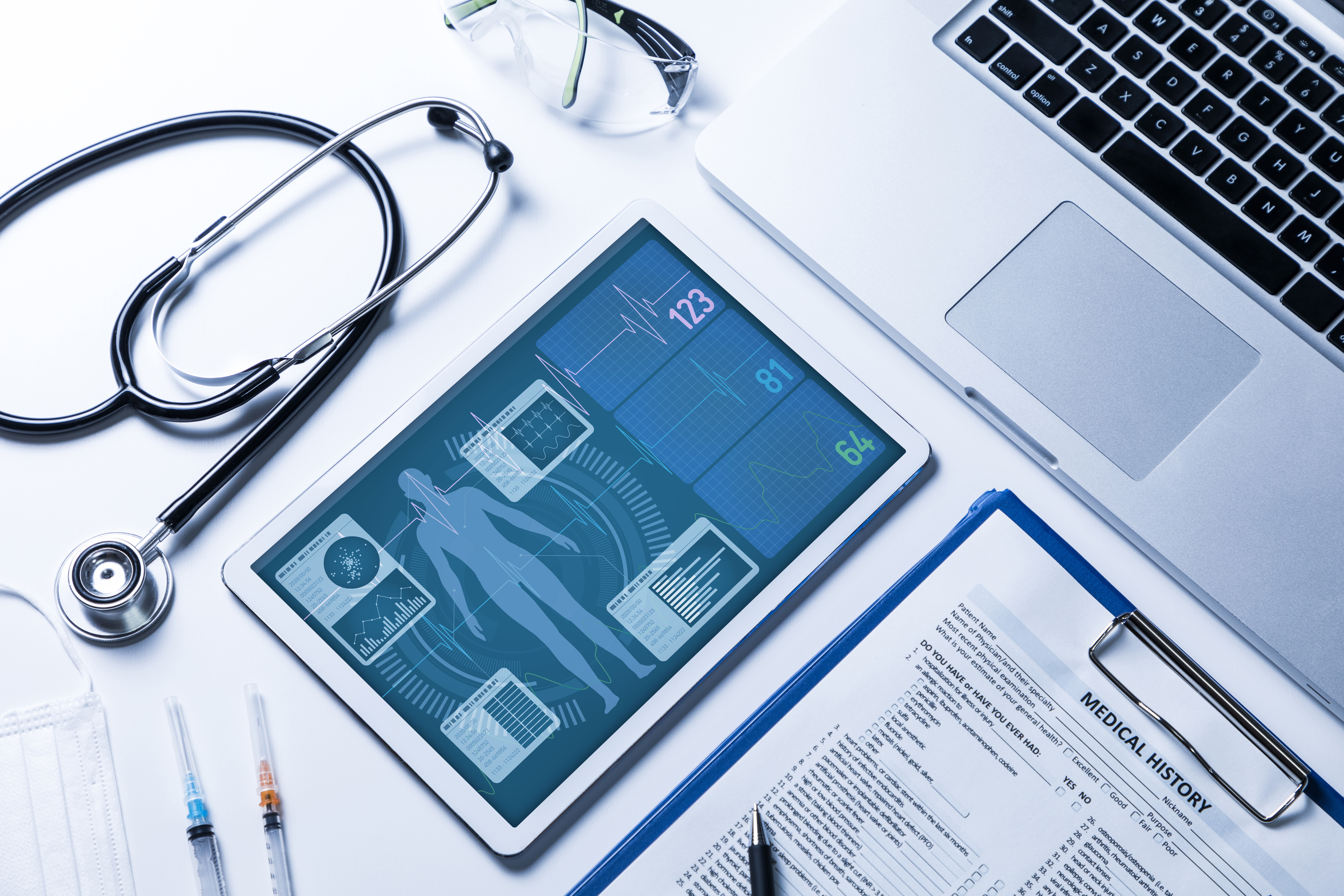 A tablet displaying vital signs and patient health data is shown alongside a stethoscope, medical syringes, a medical history form, eyeglasses, and a laptop, representing modern healthcare technology and digital patient management.