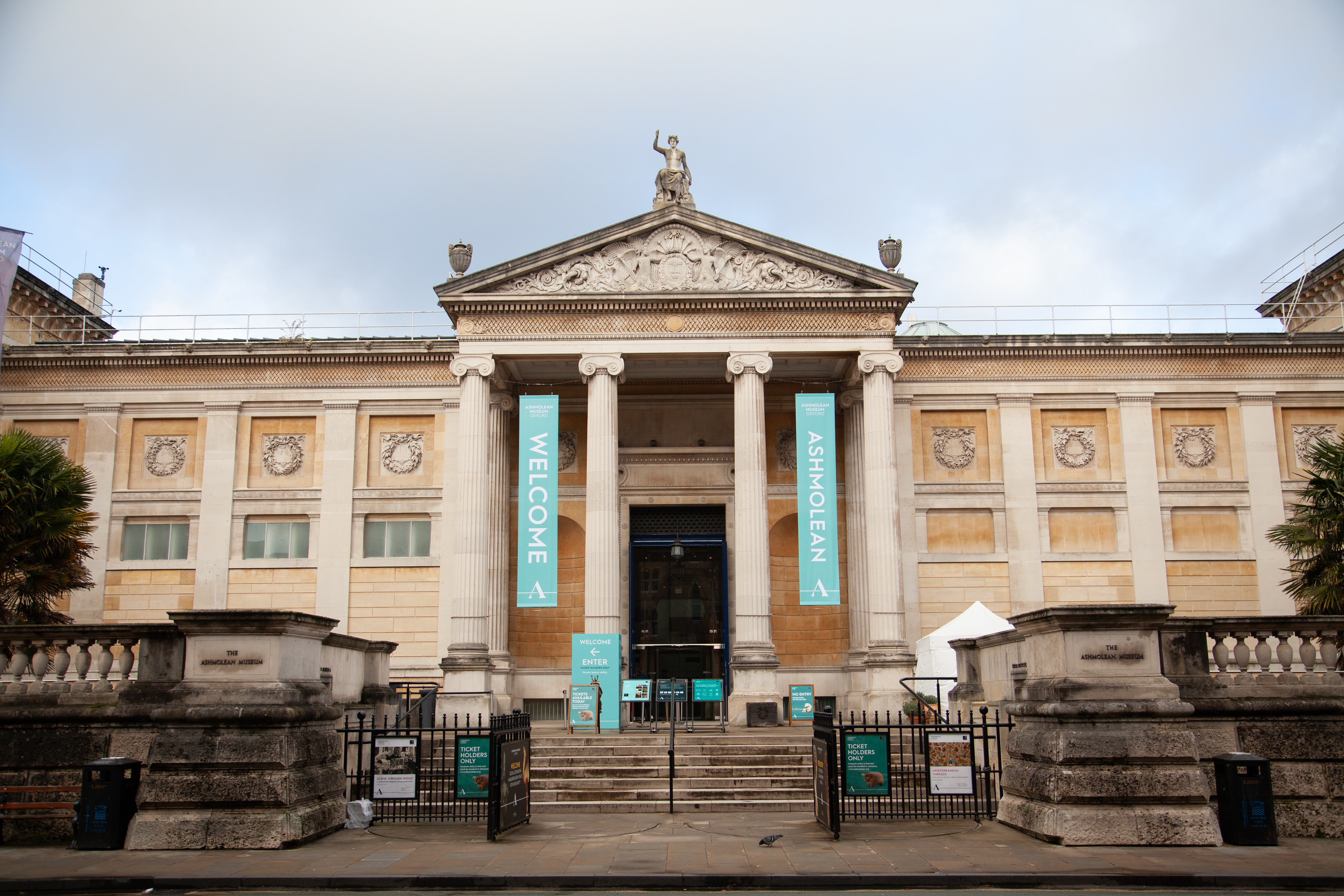The entrance to the Ashmolean Museum featuring grand columns, a statue on top, and welcome banners. The historic building is shown from the front on a cloudy day.